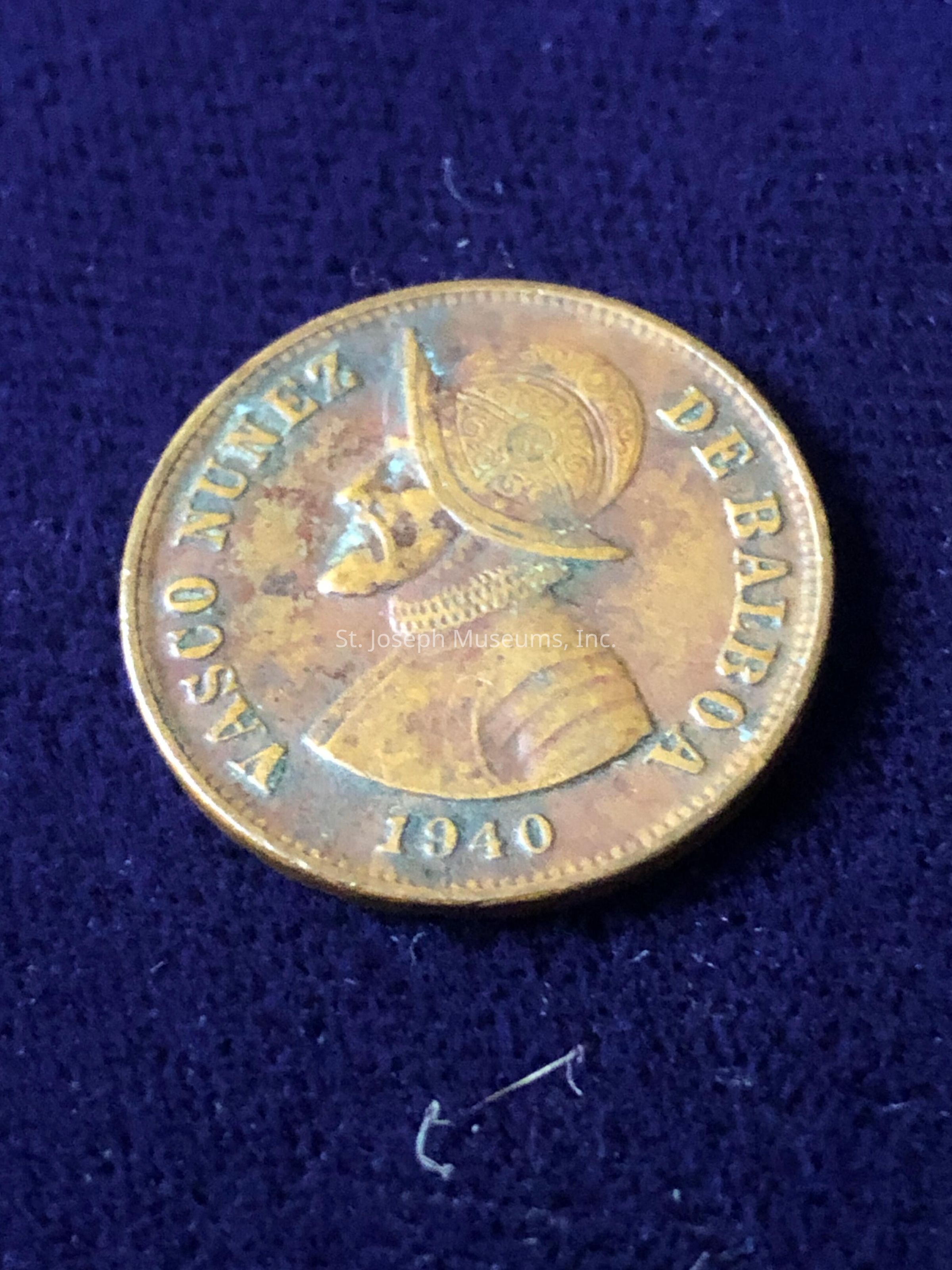 The image features a bronze-colored coin with an embossed profile of a historical figure wearing a detailed helmet. The inscription around the edge of the coin reads "VASCO NUNEZ DE BALBOA" along the top and across the bottom is the year "1940". The coin is placed on a textured, dark blue surface, which provides a stark contrast to the coin's metallic sheen. The intricate details on the helmet, including decorative patterns, are clearly visible, highlighting the craftsmanship involved in the coin's design. The coin's surface shows signs of age, with slight discoloration and tarnishing, suggesting it is an antique or collectible item. A small piece of lint is observable near the bottom edge of the coin on the fabric. The overall composition of the image draws attention to the historical and numismatic significance of the coin.