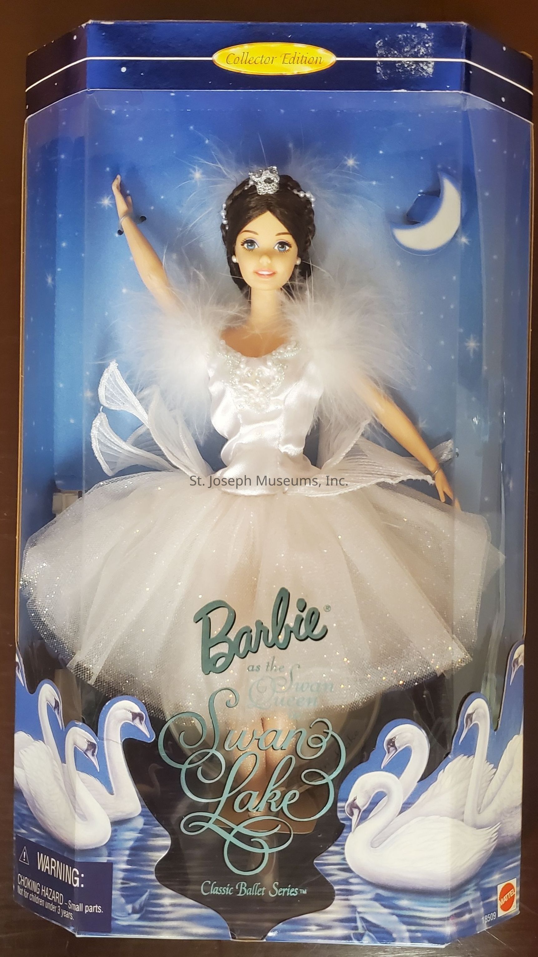 The image shows a "Collector Edition" Barbie doll from the "Classic Ballet Series," specifically styled as the Swan Queen from Swan Lake. The doll is elegantly dressed in a white ballet costume adorned with sparkling bodice details and feather-like accents on the shoulders, capturing a graceful pose with one arm raised. The background of the packaging beautifully complements the Swan Lake theme, featuring a night sky with stars and a crescent moon. The words "Barbie as the Swan Queen, Swan Lake, Classic Ballet Series" are prominently displayed on the front. At the bottom, a row of gracefully illustrated swans on a serene lake adds to the enchanting ballet motif.