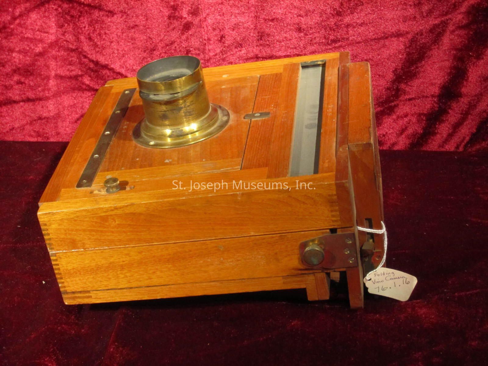 The image shows a vintage folding view camera, characterized by its wooden body and brass lens encasement. The camera is placed on a rich, velvety red surface, highlighting its classic design. The wooden construction displays a polished finish, with dovetail joints visible at the corners, indicating meticulous craftsmanship. A brass lens protrudes from the front, reflecting light from its gleaming surface. Adjacent to the camera body, a small knob and an attached metal hinge can be seen, suggesting parts of its folding mechanism. A white tags hangs at the side, inscribed with handwritten text that appears to note information about the camera. The entire scene evokes a sense of nostalgia, reminiscent of early photographic history.