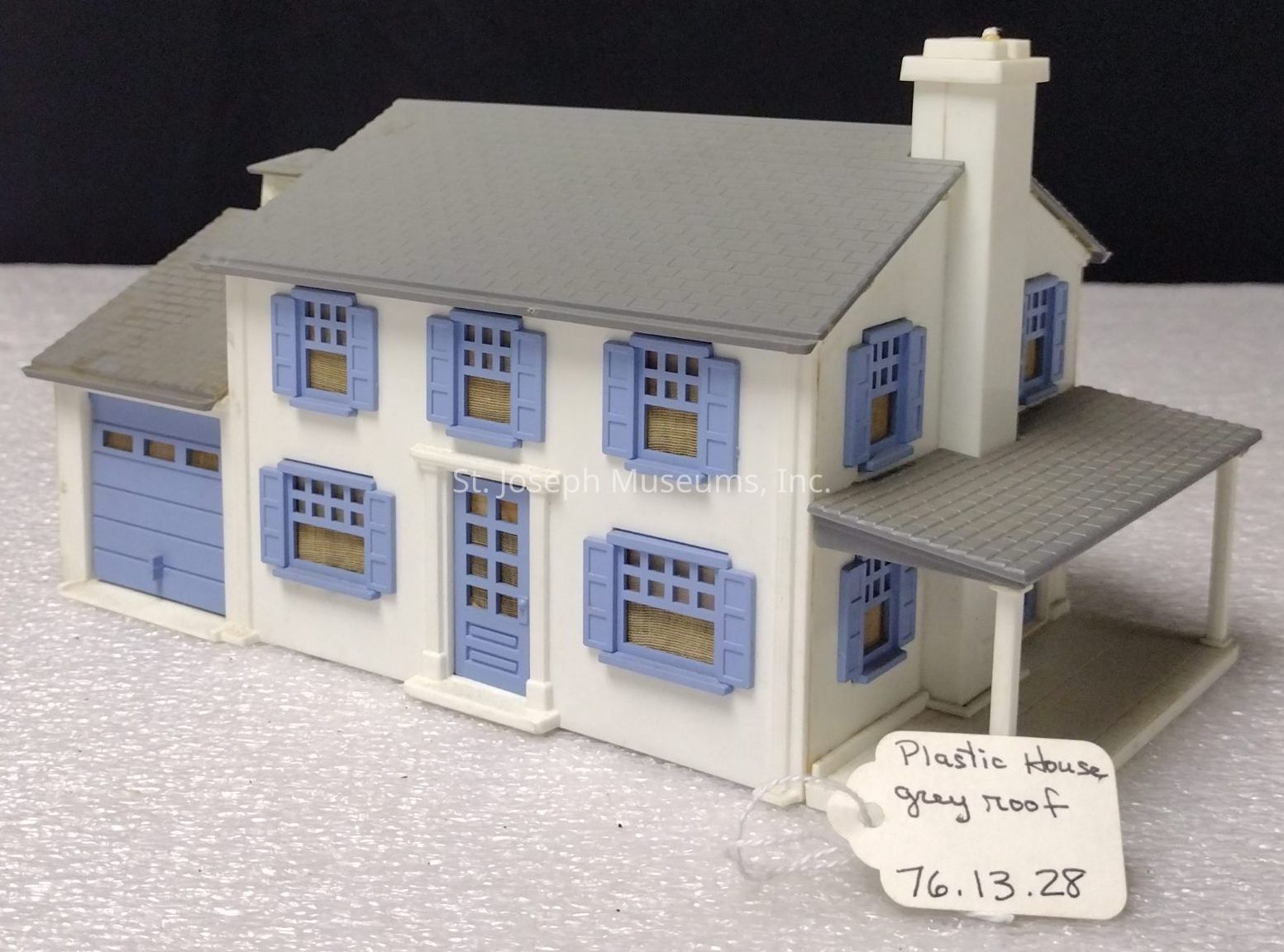 The image features a miniature plastic model house with a prominent gray roof, exhibiting a charming design that includes blue shutters and a matching garage door; the façade is mostly white, accented with delicate blue trims around the windows and door, while a small, covered porch extends from the front, supported by slender columns; the details offer a neat and stylized aesthetic, capturing the essence of traditional suburban architecture; a handwritten tag, attached to the model, reads “Plastic house grey roof 76.13.28,” providing a descriptor and potentially an identification number, all placed on a speckled surface against a plain dark background, emphasizing the miniature's quaint and composed appearance.