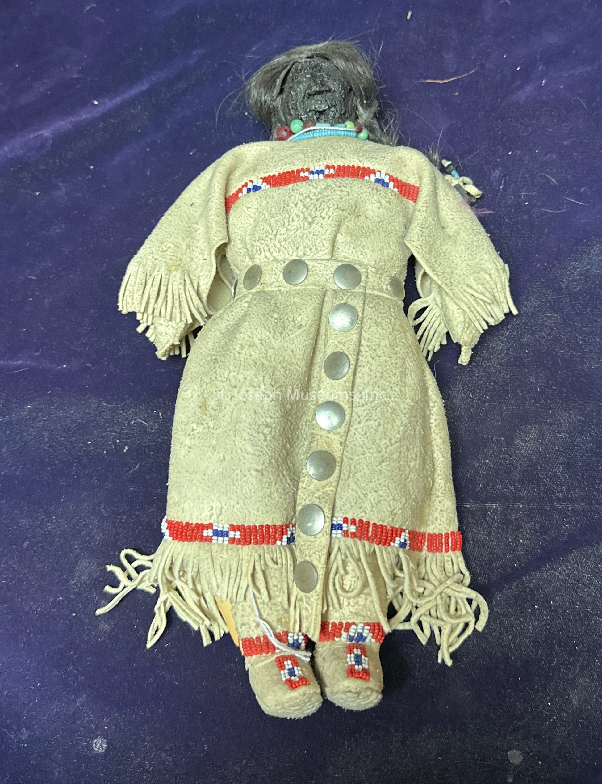 The image depicts a handmade doll lying on a purple surface. The doll wears a tan, fringed dress decorated with red, white, and blue beadwork across the chest and hem. Metal buttons run down the front of the dress, adding a decorative element. The doll's moccasins are similarly beaded, complementing the outfit. The doll's head features detailed beadwork with a necklace and has dark hair, indicating intricate craftsmanship. Overall, the doll reflects a traditional aesthetic, rich in cultural artistry, with a focus on beadwork and detailing that suggests it could be inspired by indigenous clothing styles.