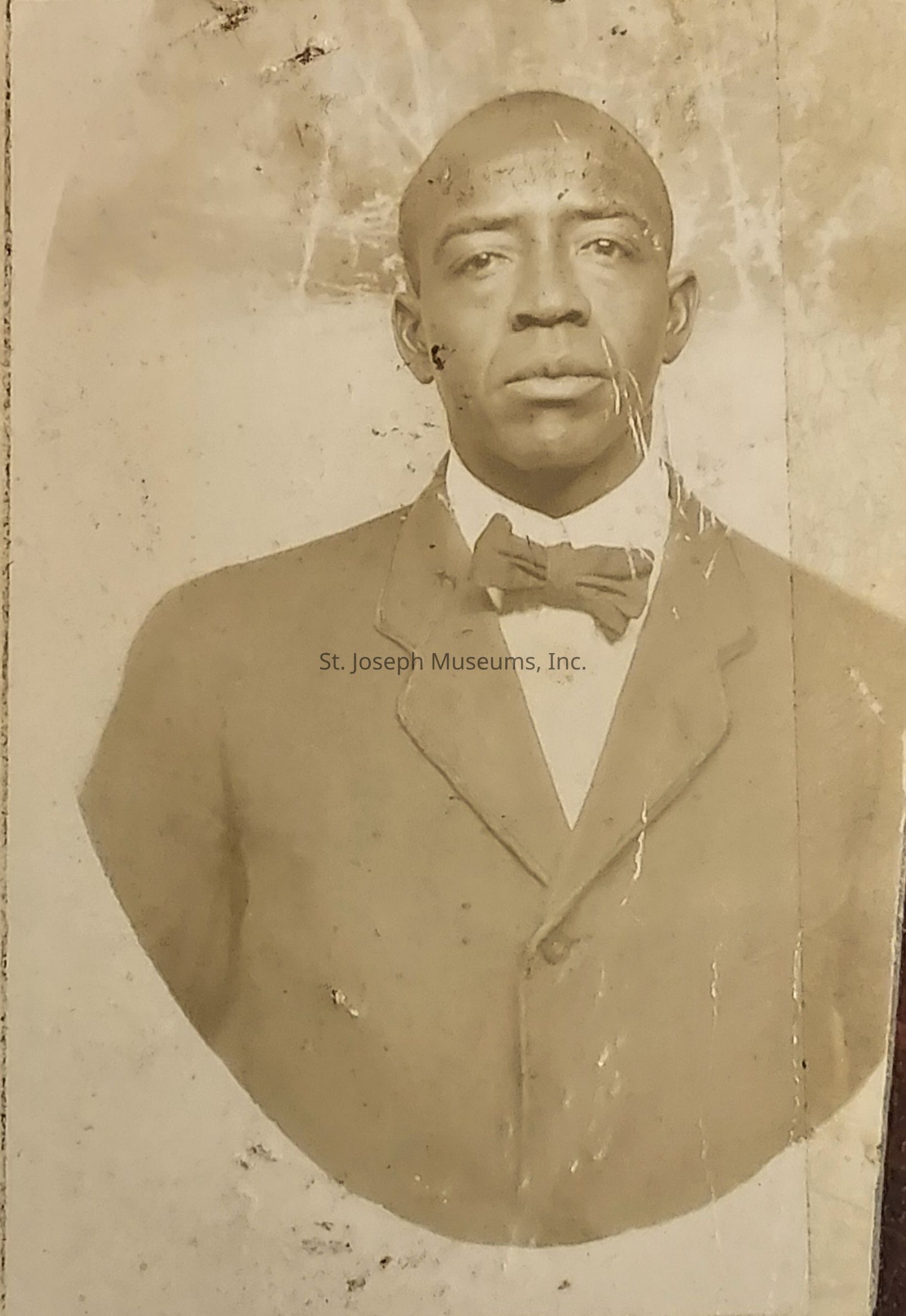 The image is an aged, sepia-toned portrait of a man captured in a formal pose, standing against a plain backdrop. He is wearing a suit with a bow tie, and his expression appears serious and composed. The photograph shows signs of wear, with visible creases and spots, suggesting it is quite old. The man’s gaze is directed at the camera, giving the portrait a straightforward and earnest quality. This historical image evokes a sense of nostalgia, capturing a moment in time with classic photography techniques.