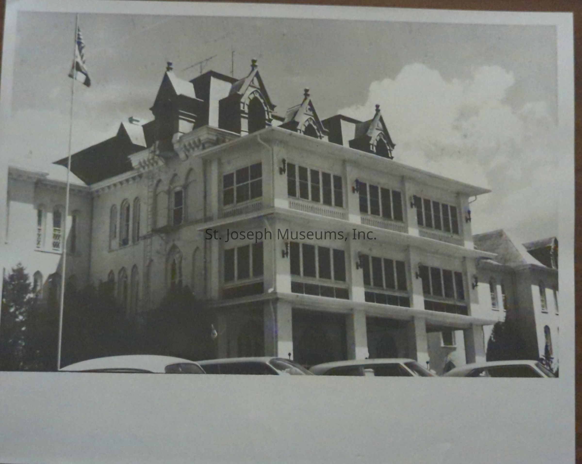Black-and-white image depicting the Missouri State Hospital Administrative Building in St. Joseph, Missouri a historic, multi-story building with ornate architectural features, large windows, and decorative roof. A flagpole stands in the foreground, indicating its institutional significance. Cars are parked in front, while fluffy clouds fill the sky, presenting a serene atmosphere.