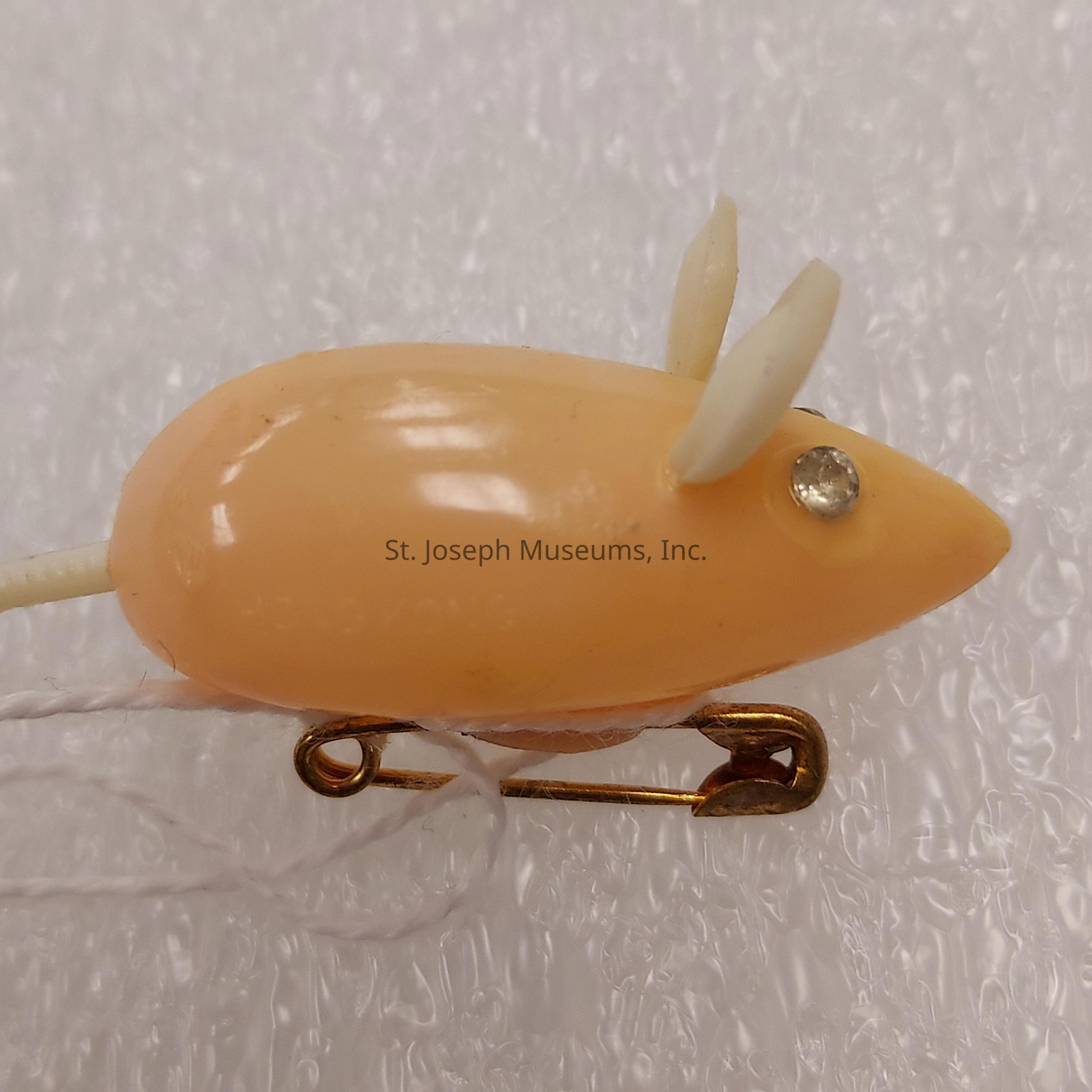 The image shows a small, cream-colored plastic object designed to resemble a mouse. It features a smooth, oval-shaped body with two small, pointed ears attached on top, as well as a shiny, rhinestone-like element representing an eye. Attached to the underside of the mouse is a metallic safety pin, likely intended for attaching the object to clothing or other surfaces. The tail appears to be made of a slender, flexible material that matches the creamy hue of the mouse's body. The background consists of a textured, white surface that highlights the pastel tones of the mouse toy, creating a soft and whimsical appearance overall.