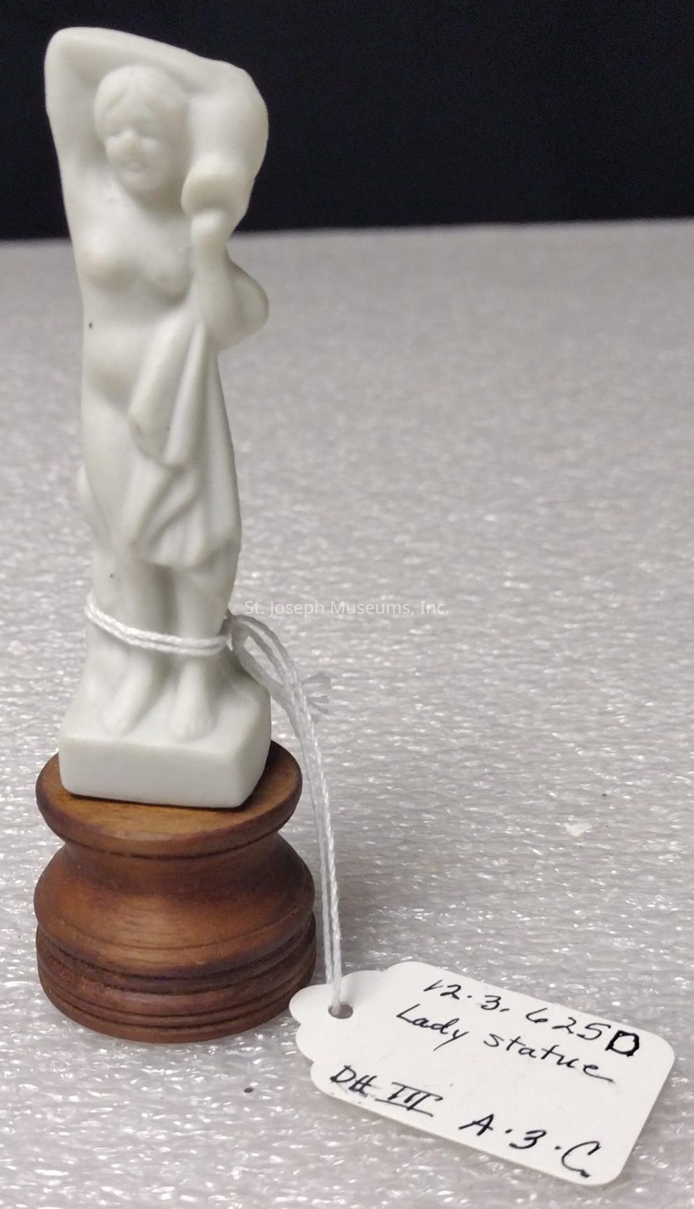 The image shows a small white statue of a lady, elegantly posed with one arm raised, crafted from what appears to be a smooth material. The figure is scantily dressed, with a cloth draped over one shoulder, enhancing its classical aesthetic. The statue is mounted on a round wooden pedestal, which is intricately turned and adds a touch of warmth through its rich brown color. Attached to the base is a small white tag secured with a thin string, containing handwritten information including numbers and the description "Lady Statue." The background is a textured surface, likely a fabric or paper, providing a simple yet effective contrast that highlights the delicate details of the sculpture.
