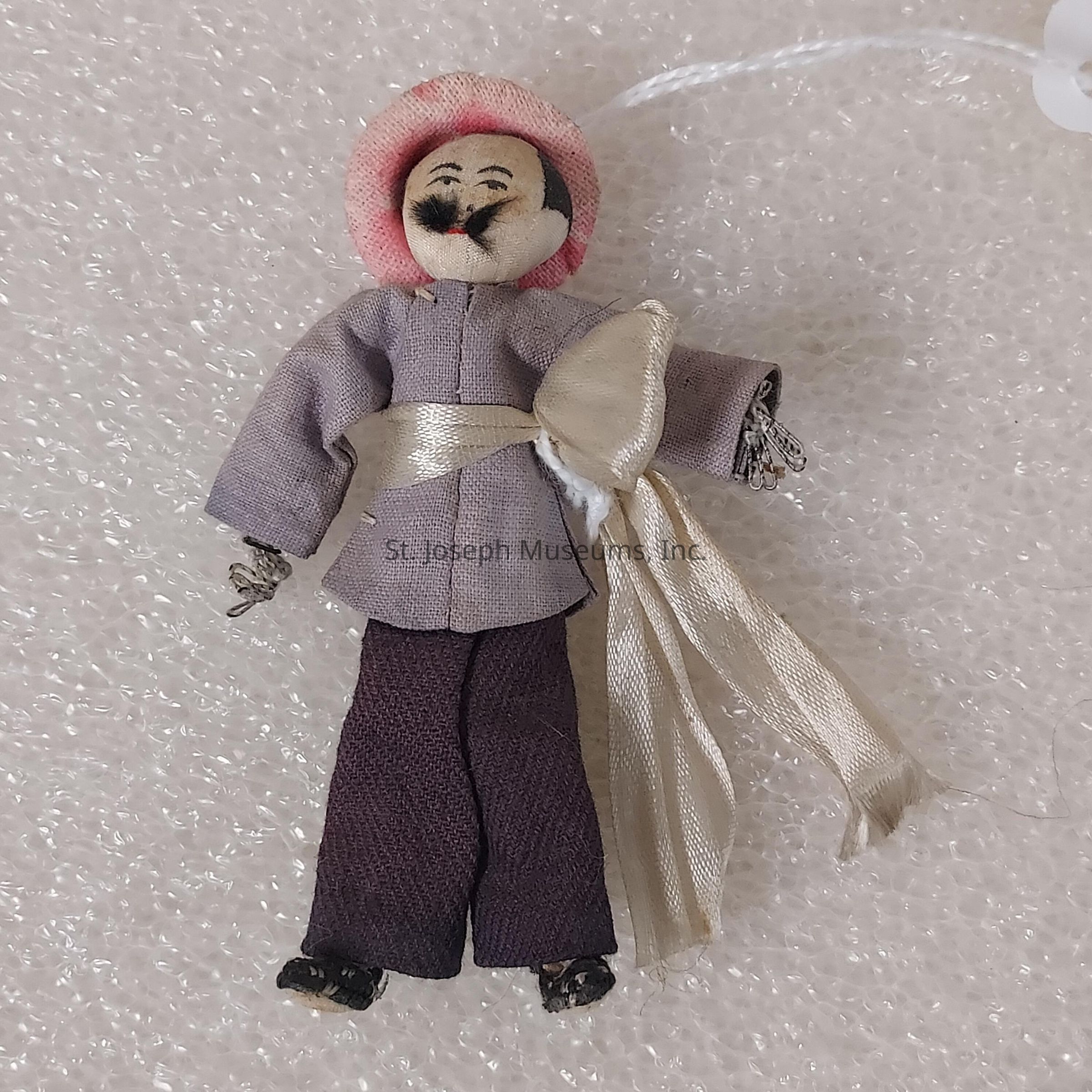 The image shows a small handcrafted doll featuring a detailed face with painted features, including eyes, eyebrows, and a mustache. The doll is dressed in a traditional outfit consisting of a light gray top tied with a cream-colored sash and dark pants. It wears a pink headpiece that adds a pop of color to its ensemble. Its hands appear intricately made, and the feet are detailed to look like shoes. The doll is set against a textured background that resembles a bubble wrap surface, which provides a soft and neutral backdrop to highlight the doll's craftsmanship and attire. Overall, the image captures the charm and artistry of the doll's design, showcasing the vibrancy and cultural aspects of its clothing and facial expression. The attention to detail, from the fabric choices to the facial painting, indicates a handcrafted piece likely representing cultural or folkloric significance, providing insight into traditional attire or artistic expression from its place of origin.