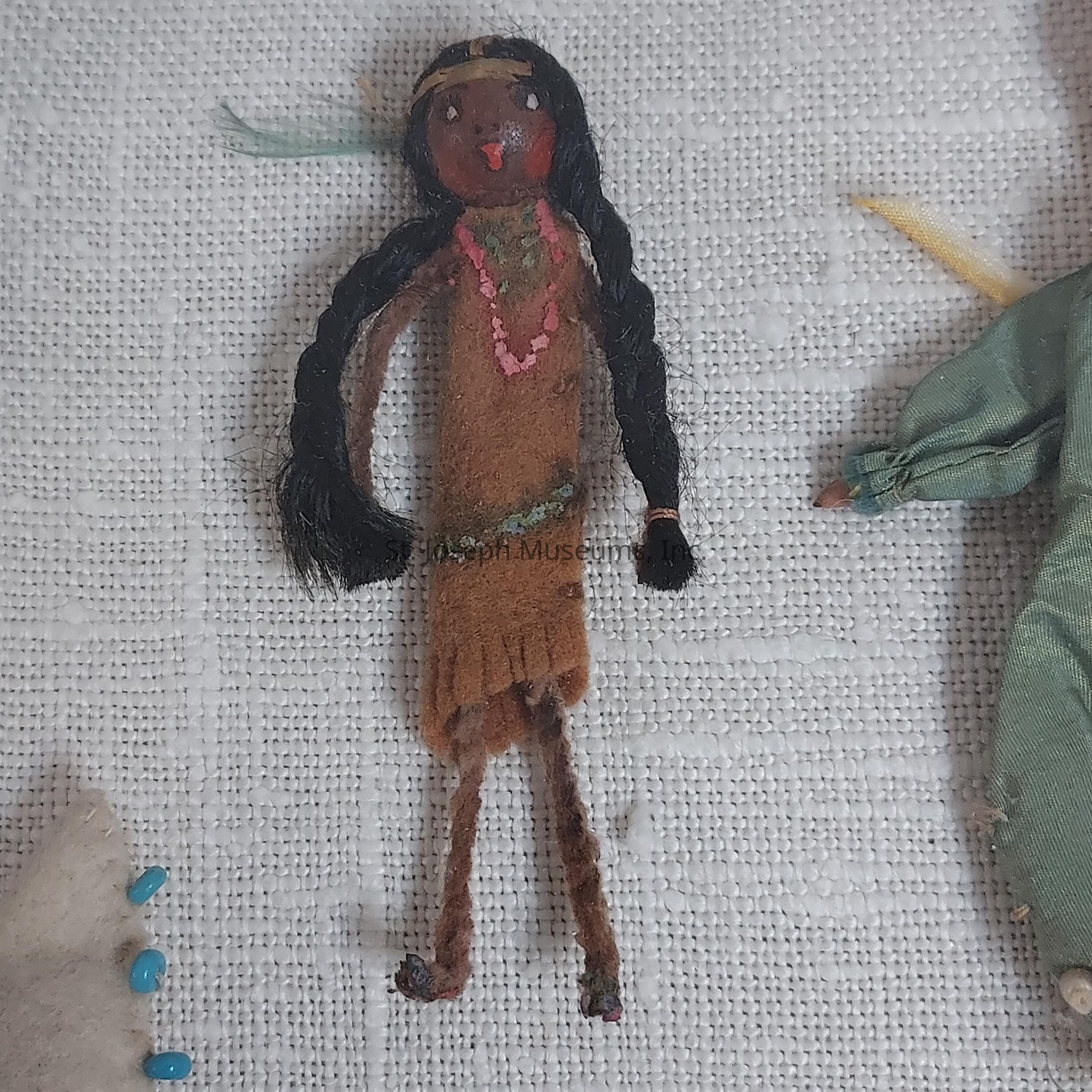 The image depicts a small, handmade doll crafted from fabric and yarn, positioned on a textured, light-colored background. The doll has long black yarn hair styled into braids, a headband, and a beaded necklace. It wears a felt dress in earthy tones of brown with fringe details. The facial features are expressive, with painted eyes and lips adding character. The arms and legs are slender and extended in a playful manner. This doll likely reflects a cultural or historical motif, as suggested by its attire and adornments. It is part of a collection, suggested by the partial view of other similar objects on either side.
