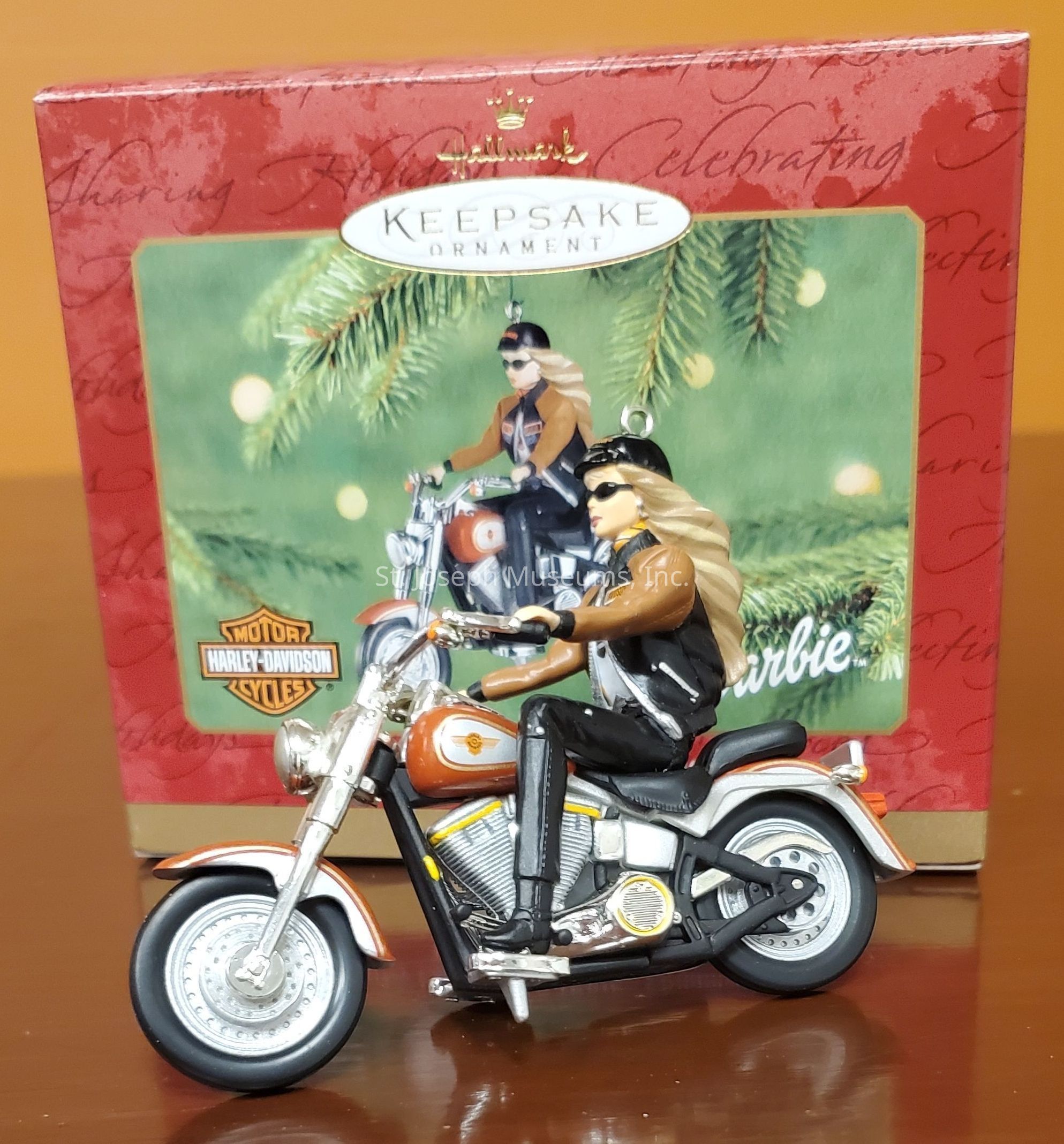 The image features a Hallmark Keepsake ornament depicting a figure on a Harley-Davidson motorcycle. The ornament showcases a person with long blonde hair, wearing a brown jacket, black pants, and sunglasses, embodying a biker aesthetic. The Harley-Davidson logo is visible, enhancing the iconic motorcycle theme. The background box displays festive elements with pine branches, and the ornament inside mirrors this design. The combination of holiday decor and biker culture creates a unique collectible item, appealing to enthusiasts of both motorcycles and festive decorations. The overall presentation highlights the blend of adventure and celebration.