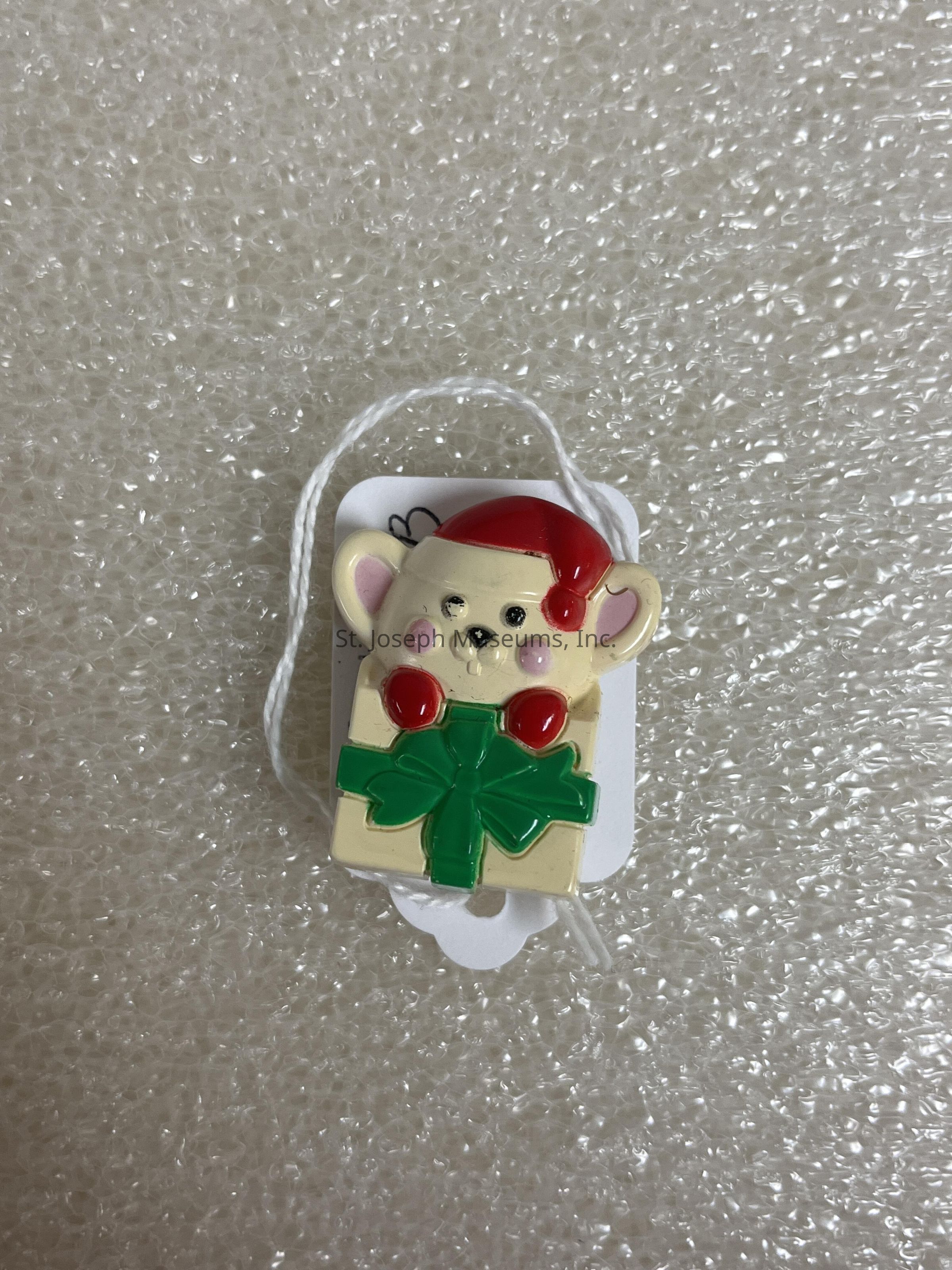 The image depicts a cute, festive ornament featuring a small, cartoon-like mouse character that has large ears and a cheerful expression, adorned in a red Santa hat with a white pom-pom and a small red dot detail. The mouse is also sporting blushing cheeks and is accompanied by a pair of bright red berries just below its face. To add to the festive flair, the character is attached to a yellow square with an exaggerated, shiny green bow placed at the center, symbolizing a wrapped Christmas present. This charming ornament is set against a textured background made of bubble wrap, adding an interesting contrast to the scene. Accompanying the ornament is a white string looped to a tag for easy hanging. The tag peeks slightly from behind the ornament, hinting at a decorative purpose, possibly to be hung on a Christmas tree, wreath, or other holiday display.