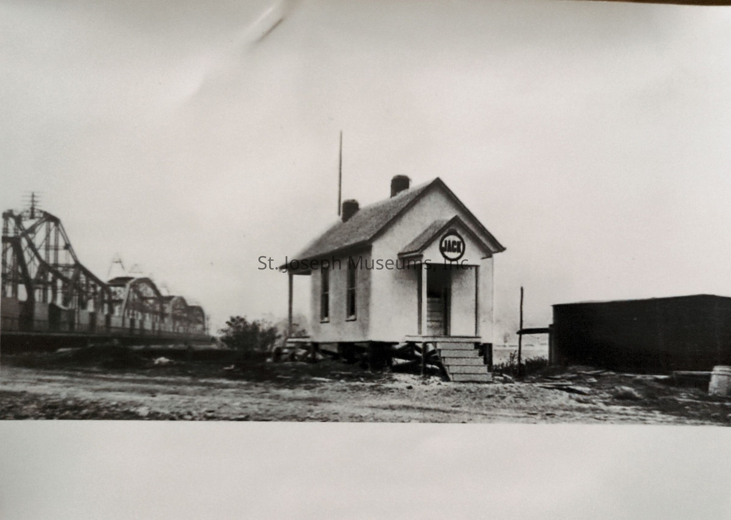 Black-and-white image depicting a small office or house on supports, with a sign above the entrance, positioned near a bridge spanning the Missouri River, crossing into Kansas. The bridge features multiple arches and trusses, while the building appears modest and isolated, surrounded by an empty, barren landscape.