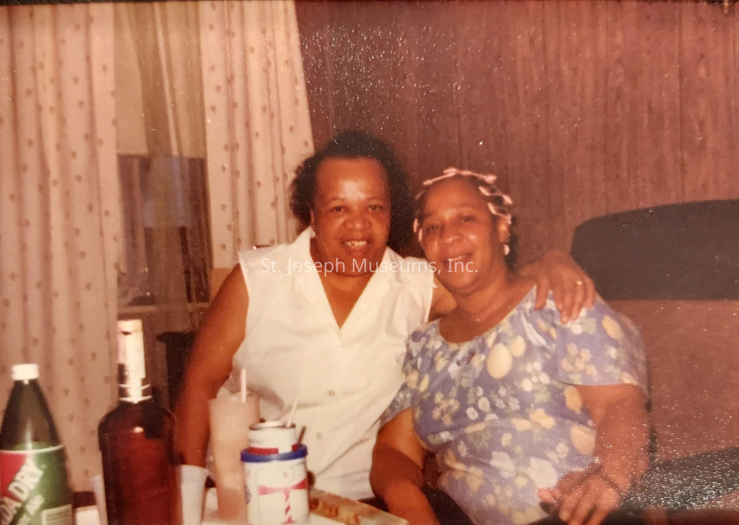 The image shows two women sitting closely together in a room with wooden panel walls and polka dot curtains. They are smiling at the camera, creating a warm and friendly atmosphere. One woman is wearing a white sleeveless top, while the other is wearing a blue floral blouse. They have their arms around each other, suggesting a close relationship. In front of them on the table are various items, including bottles and cups, indicating they might be enjoying a casual drink together. The scene conveys a sense of companionship and comfort.