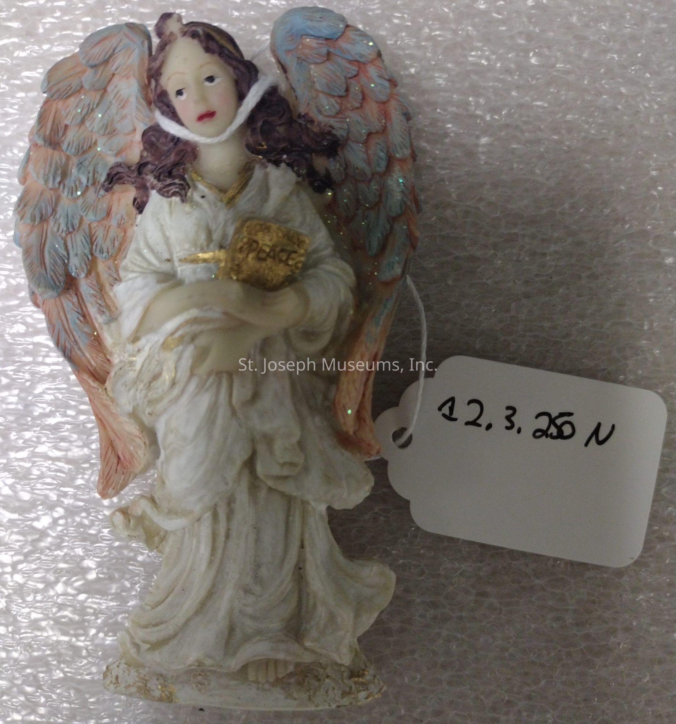 The image features a delicate figurine of an angel with intricately detailed wings, painted in shades of blue and pink, holding a golden object inscribed with the word "Peace." The angel, robed in flowing white garments, has gently curled hair and a serene expression. The figurine stands out against a textured surface beneath, and a small white tag is attached to the angel with a string, bearing a handwritten notation: "12,3,25 n." The piece emanates an aura of tranquility and artistic craftsmanship, evoking a sense of gentle grace and celestial beauty, making it a fitting ornament for holiday or spiritual decor.