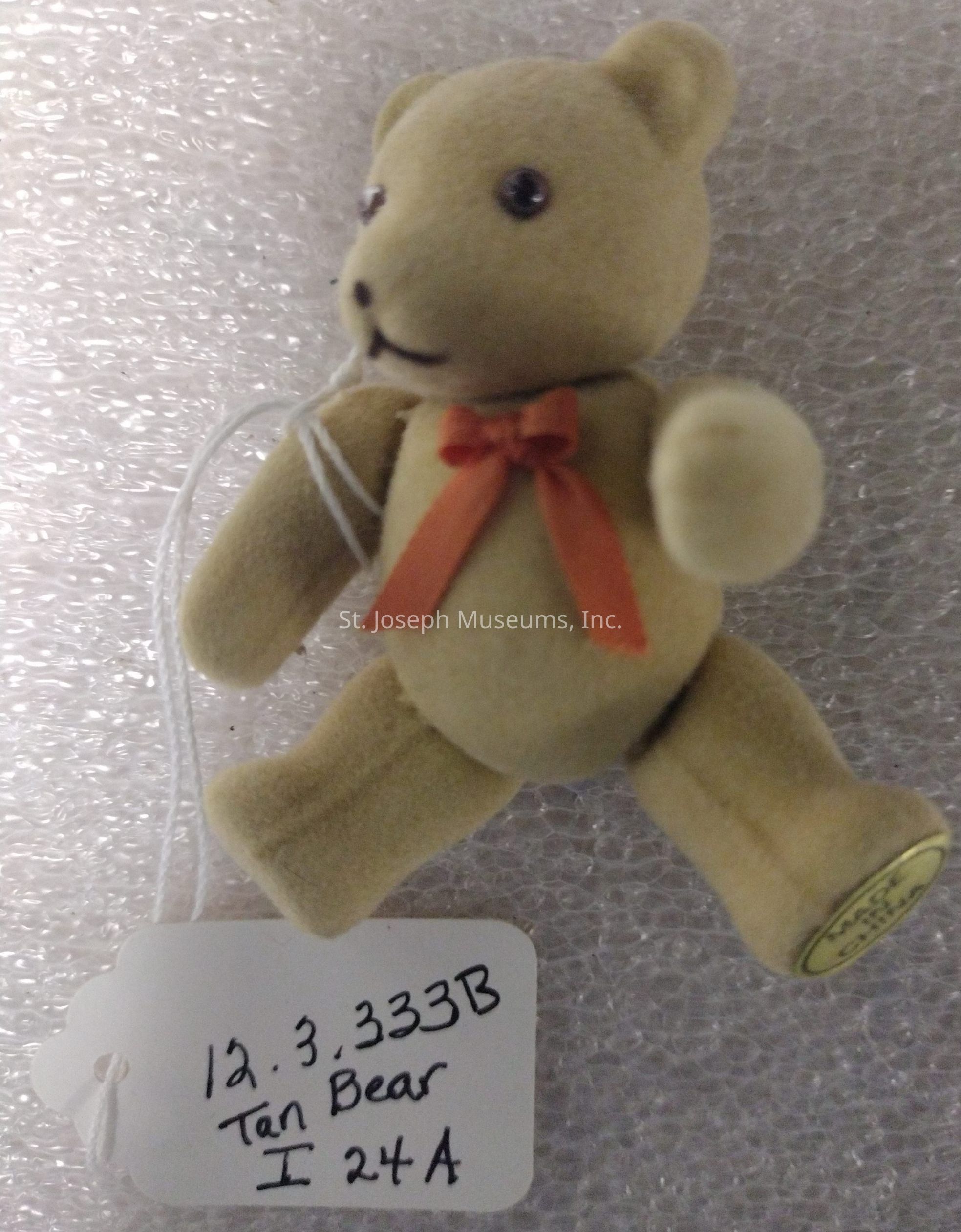 The image features a small tan teddy bear with a soft, plush texture, adorned with a bright orange bow around its neck, giving it a charming appearance. The bear has shiny black eyes and a stitched black nose and mouth, adding to its cute and classic teddy bear look. Attached to the bear with a string is a white rectangular tag with handwritten information: "12.3.333B, Tan Bear, I24A." The background consists of a textured surface that resembles bubble wrap, likely used for protective packaging. The tag’s details indicate some sort of cataloging or identification system, possibly for inventory or collection purposes, suggesting that the bear might be part of a larger set. The overall presentation of the bear, with its classic design elements and attached tag, conveys a sense of nostalgia and care, highlighting its status as a collectible or special item.