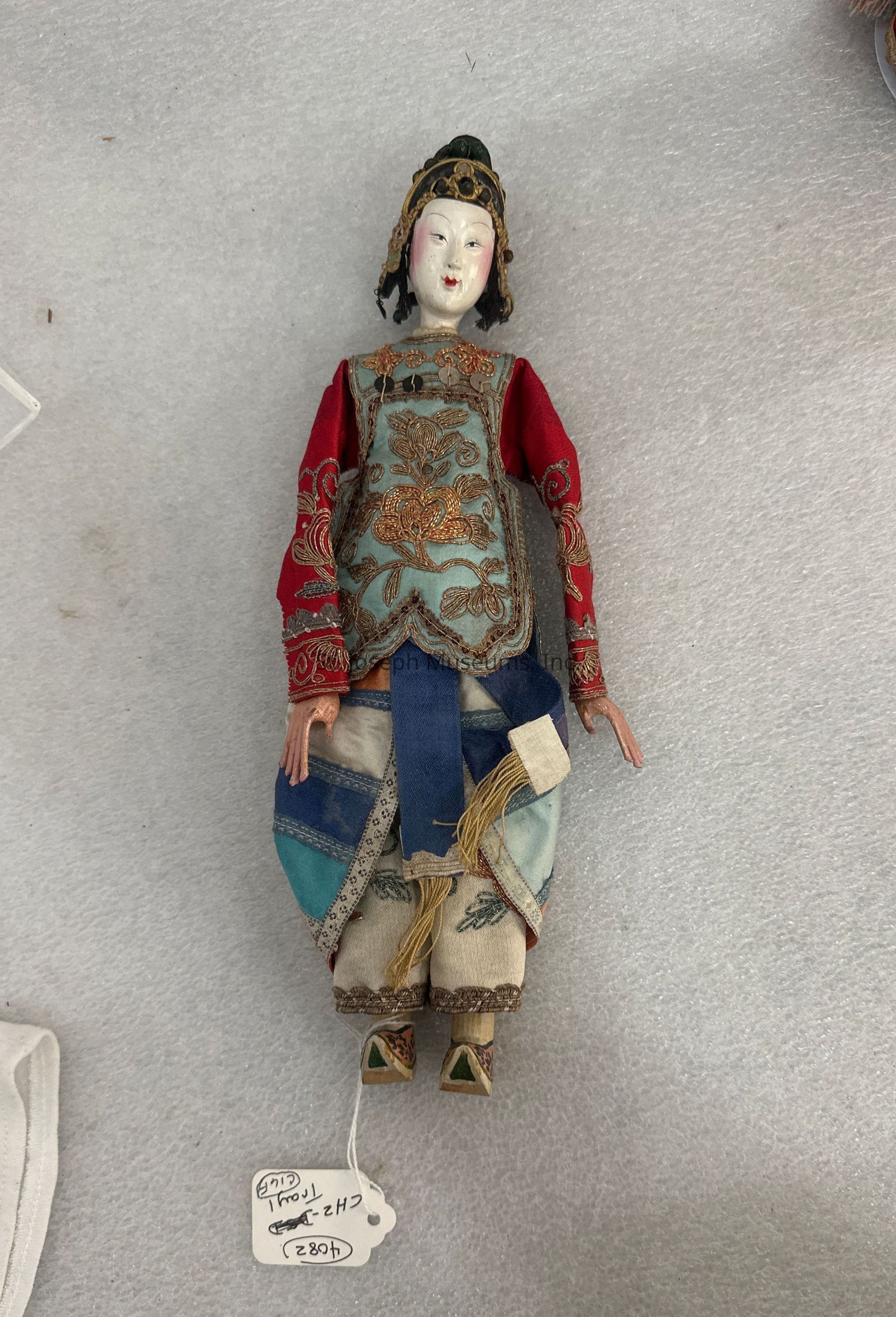 The image features a traditional doll with a beautifully crafted face and ornate attire, lying on a textured surface. The doll wears an elaborate outfit consisting of a red and blue brocade top adorned with gold floral embroidery and intricate patterns. The sleeves are red and match the top's vibrant embroidery, while the lower half of the attire consists of baggy pants made from patchwork fabric in shades of blue and cream. Accessories include a headpiece with a dark hue and metallic embellishments, complementing the overall design. The doll has painted features with distinct eyes and lips, and its articulated limbs add to its detailed craftsmanship. At the bottom of the doll, small shoes feature a triangular green detail. A white tag with handwritten text is attached, indicating its classification or description, enhancing the doll's aesthetic and showcasing its cultural significance. Overall, the image portrays a piece of artistry that reflects cultural heritage and meticulous attention to detail in its creation.