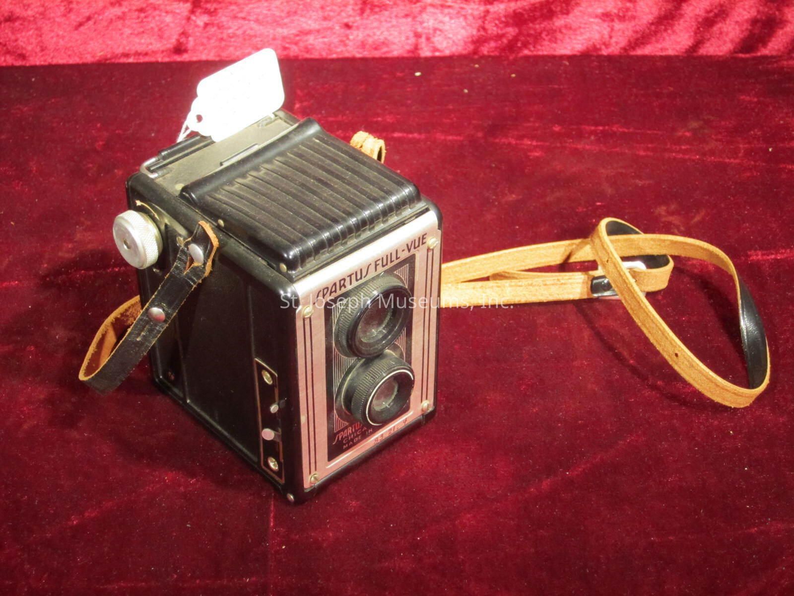 The image showcases a vintage camera labeled "Spartus Full-Vue," resting on a rich red velvet surface. The camera features a classic box design with dual lenses, a textured top panel, and a side-mounted winding knob. A tan leather strap is attached, providing a rustic touch to the camera’s appearance. The surface on which it rests adds a luxurious feel, enhancing the camera's antique charm. A small white tag is attached to the top, possibly indicating a price or catalog information. The overall composition highlights the craftsmanship and nostalgic appeal of the vintage photographic equipment.