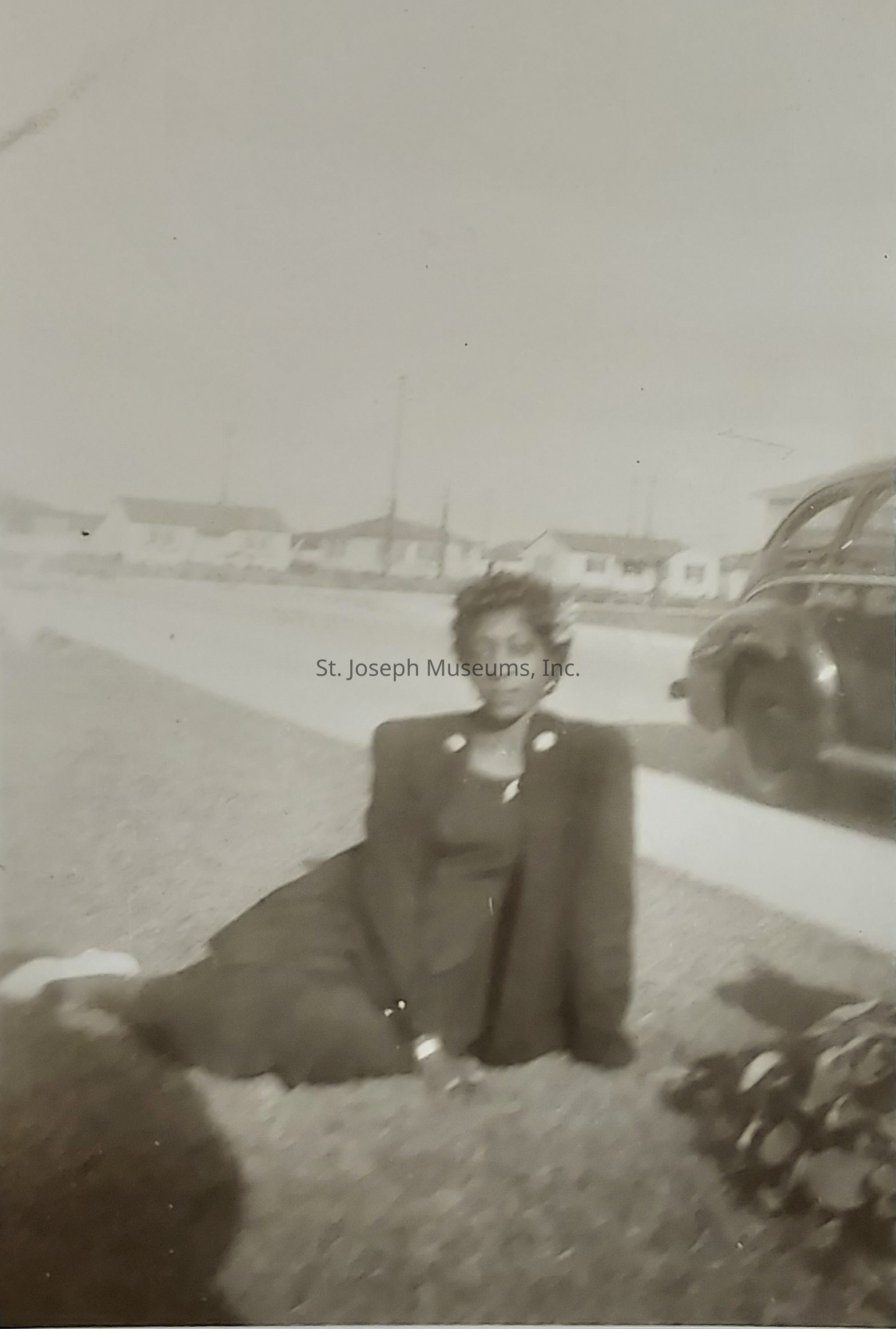 In this vintage black-and-white photograph, a woman is sitting on the grass with her legs extended towards the camera, dressed in a formal dark outfit, possibly a coat with statement buttons, and appears to be posing naturally while looking ahead; the background features a row of small houses lined along a street, indicating a suburban neighborhood, while an old-fashioned car is parked on the road to the right, evoking a sense of nostalgia and capturing a moment from the past that speaks to the fashion and lifestyle of that era, with soft focus adding a dreamy quality.