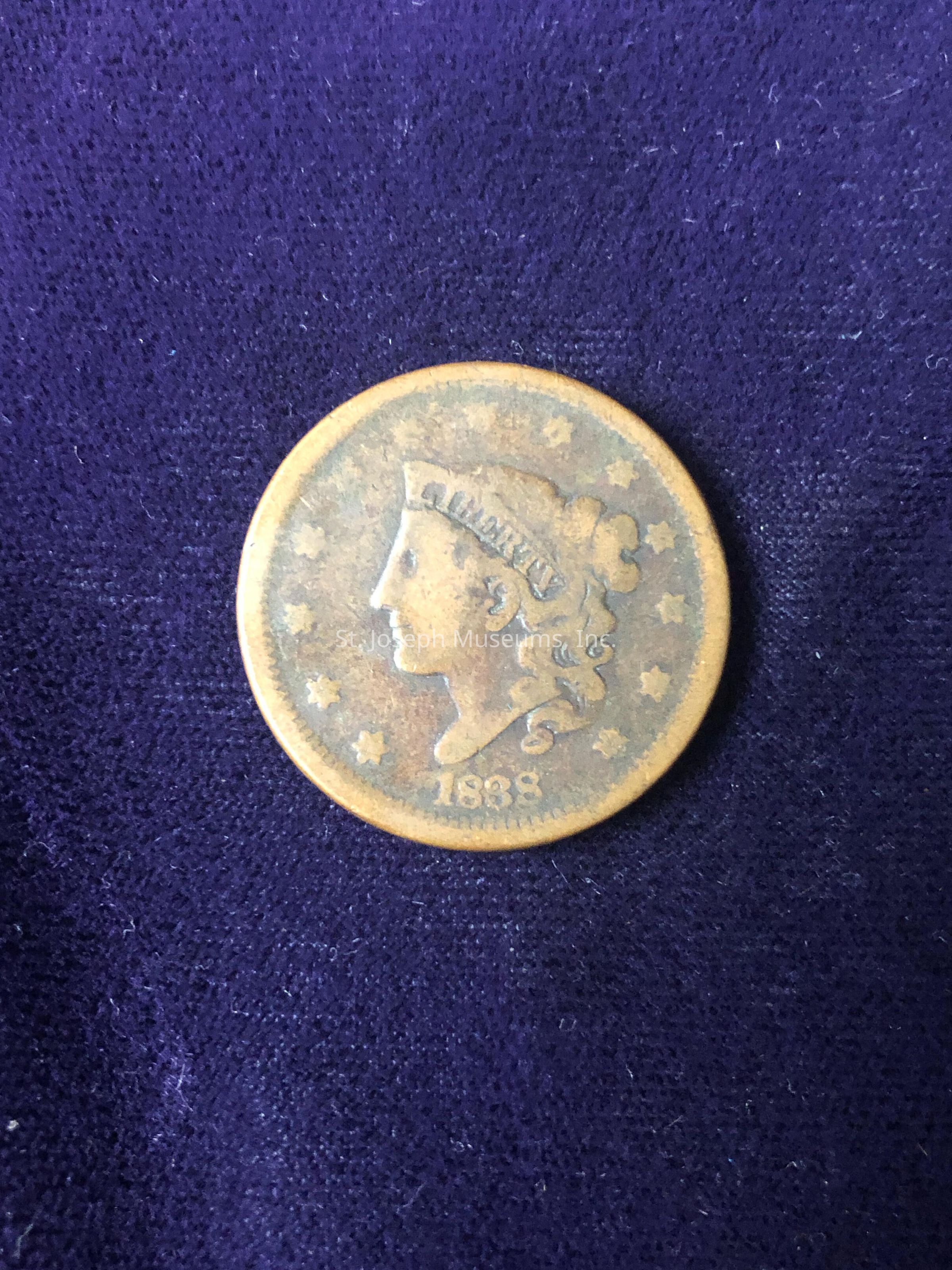 The image displays an antique coin dated 1838. The coin features a side portrait of a woman facing left, adorned with a headband inscribed with an indecipherable text. Surrounding the portrait is a ring of stars, adding to the ornate design typical of coins from that era. The coin's hue appears weathered, indicating its age and circulation over the years, with shades of brown and patina giving it a rustic look. The details on the coin suggest it may be a representation of a historical figure, possibly indicative of the country's currency during that period. The interplay between the coin's metallic sheen and the plush purple background creates a visual contrast, highlighting the coin as the focal point of the image. This juxtaposition emphasizes the coin's historical and potentially numismatic value, appealing to collectors and history enthusiasts. The overall composition captures a moment of numismatic nostalgia, celebrating the intricate craftsmanship of 19th-century minting techniques.