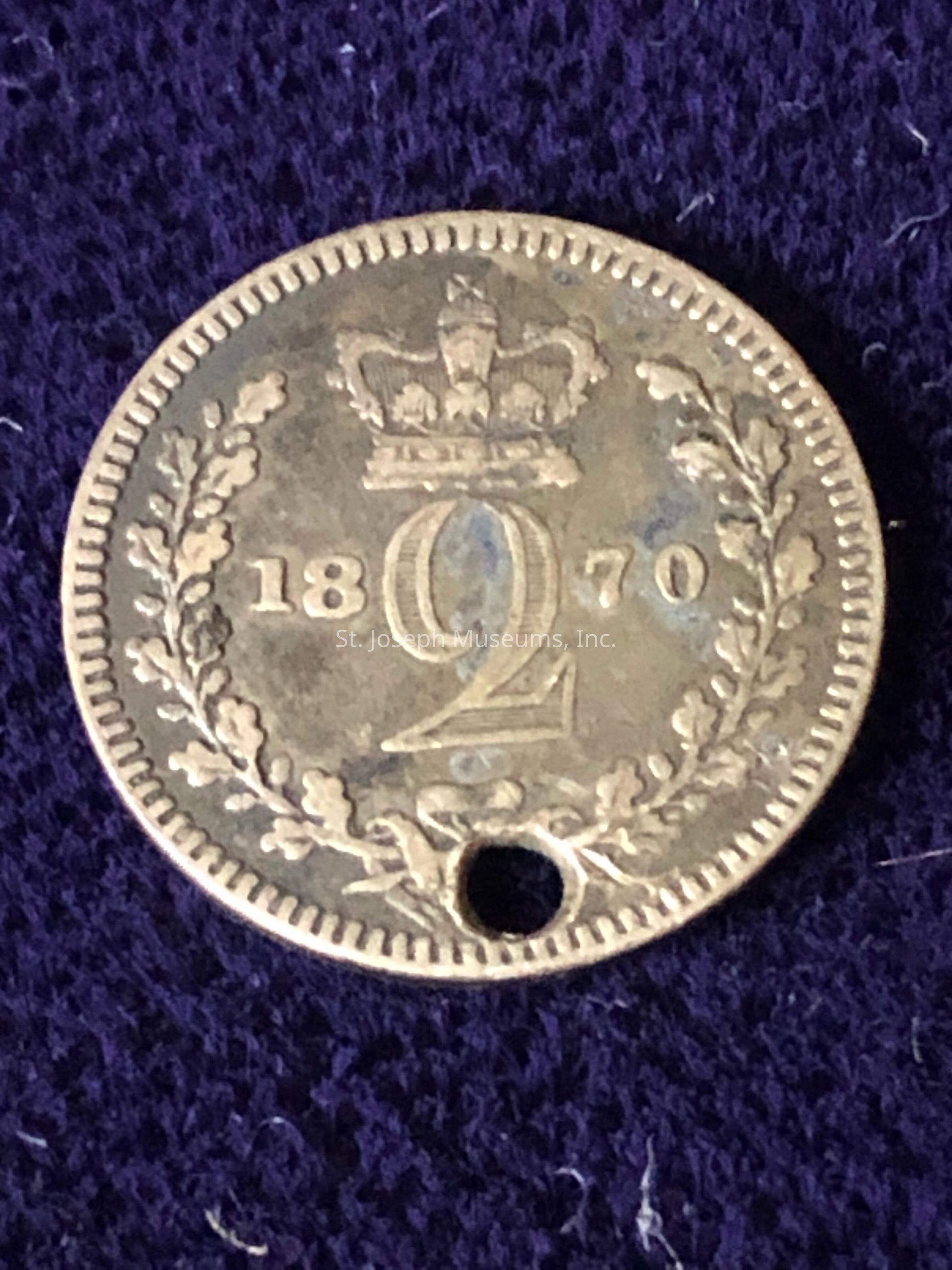 The image depicts an antique coin from the year 1870, featuring a denomination of "2" prominently embossed in the center, surrounded by decorative elements. At the top of the coin is an intricate crown design, symbolizing its likely historical and royal connection. The year "1870" is split around the central number, with "18" on the left and "70" on the right. Encircling the number is a laurel wreath with stylized leaves, which adds elegance and regal symbolism to the piece. The edge of the coin is ridged, providing an additional layer of decorative detail. Notably, there is a small hole at the bottom edge, suggesting that it may have been used as a charm or pendant at some point. The coin rests on a dark, possibly fabric, background that highlights its worn metallic texture and historic character.