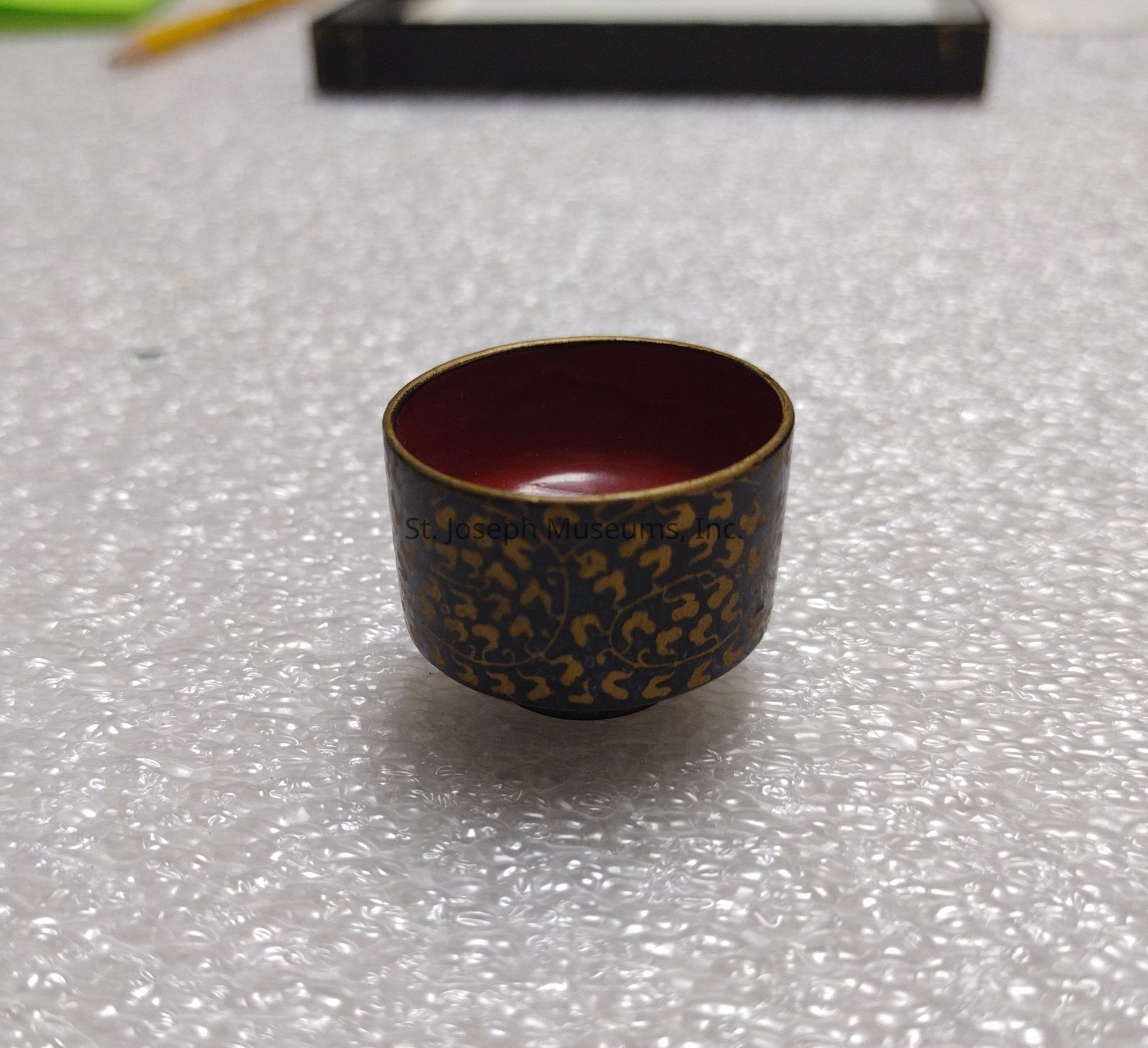 In the image, there is a small cup placed prominently in the center, characterized by its intricate and elegant design. The exterior of the cup features a dark, possibly black or deep brown surface adorned with golden patterns, which appear to be ornate and possibly inspired by traditional motifs. These patterns might suggest a cultural or historical influence, lending an air of sophistication to the object. The interior of the cup is a rich maroon or burgundy color, which contrasts beautifully with the exterior and adds depth to the piece. The cup is standing on a surface covered with bubble wrap, indicating that it might be in a storage or packaging setting, possibly for protection or display. In the background, there is a pencil and a rectangular object, maybe a box, which might suggest a workspace or a setting where items are being organized or cataloged. The image as a whole conveys a sense of careful preservation and appreciation for the craftsmanship represented by the cup.