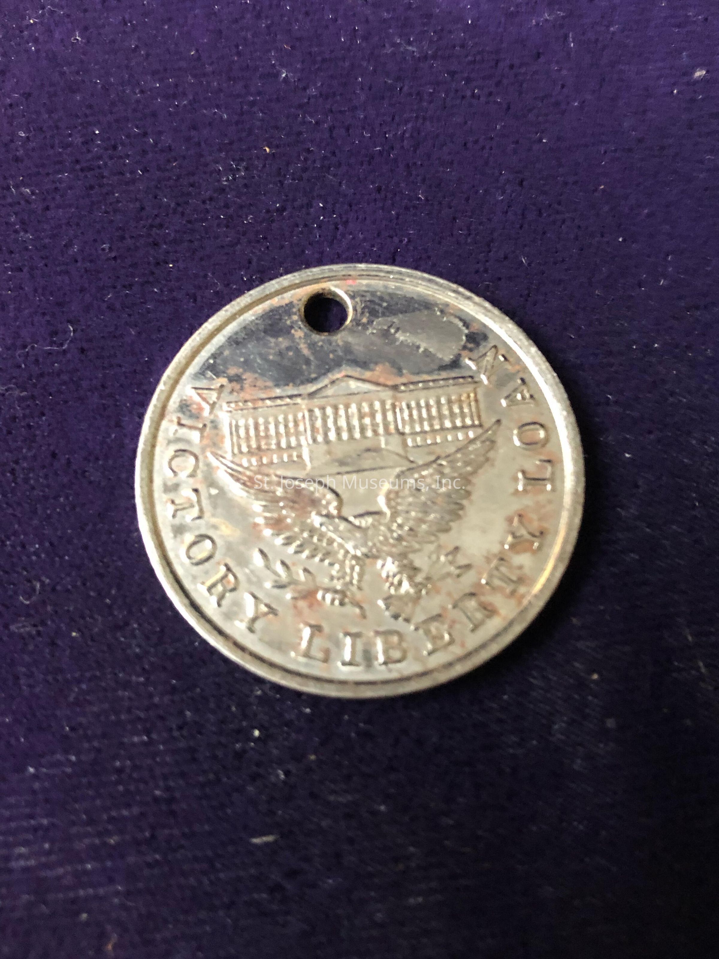 The image features a circular metallic medallion with a small hole near the top edge, suggestive of its use as a pendant or charm. The surface of the medallion is engraved with a depiction of an eagle, symbolizing strength and freedom, holding a cluster of arrows and an olive branch in its talons, representing the concepts of war and peace. Above the eagle is an image resembling a classical building with pillars, possibly symbolizing government or democracy. Encircling these motifs is the inscription "VICTORY LIBERTY," reflecting themes of triumph and freedom.