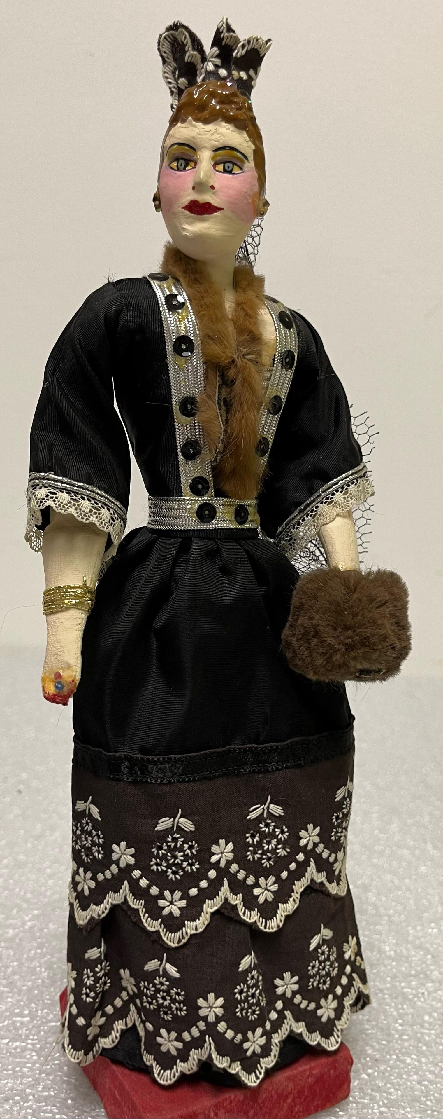 The image displays a doll dressed in an elegant, vintage outfit. The doll is wearing a black dress adorned with intricate lace and floral embroidery, featuring scalloped edges. Its sleeves are detailed with lace trim, and the bodice is decorated with silver accents and circular embellishments. Around the neck, there is a fur trim that matches the small fur muff the doll is holding in one hand. The doll has a painted face with bright red lips and pink cheeks, along with detailed eyes and eyebrows. Its hair is styled with a decorative black and white headpiece. The arm has a bracelet, adding to the overall ornate appearance. Additionally, the doll stands on a red platform against a neutral background.