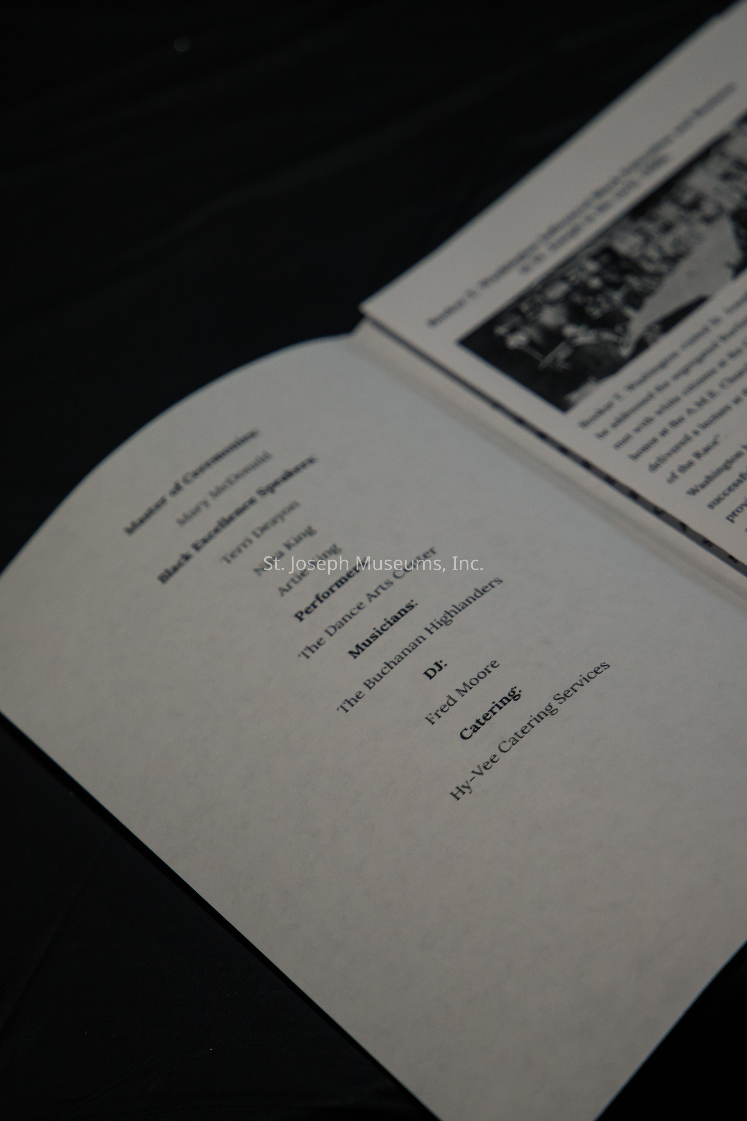 The image showcases an open event program on a dark surface, listing various contributors and participants involved in the event. On one page, the text includes categories such as "Mistress of Ceremonies," with a name underneath, "Black Excellance Speakers," listing three individuals, and "Performers," naming "The Dance Arts Center." The program also details "Musicians," specifically mentioning "The Buchanan Highlanders," followed by a section for the "DJ," naming "Fred Moore," and finally, "Catering," highlighting "Hy-Vee Catering Services." The adjacent page contains text and a blurred black-and-white image, suggesting additional event information or a historical reference. The program suggests a well-organized event, emphasizing cultural celebration and professional involvement.
