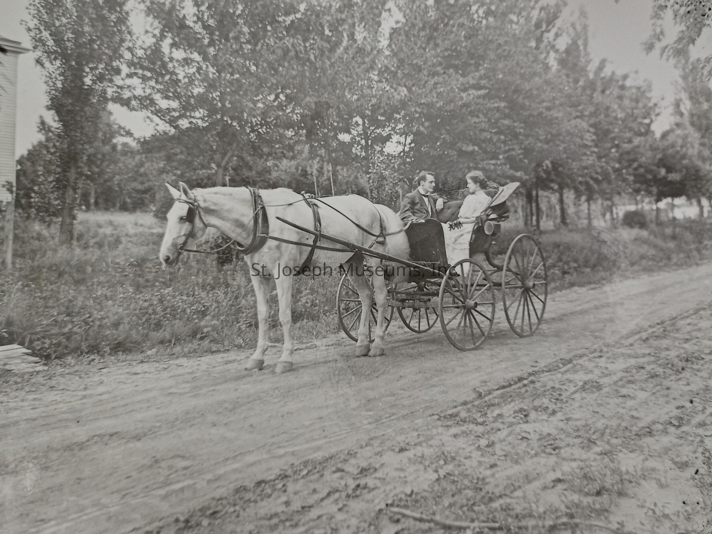 Black-and-white photograph of a couple enjoying a leisurely ride in a horse-drawn buggy along a serene country road known as Lovers Lane, surrounded by lush trees under a clear sky. The man and woman appear comfortably seated, engaged in conversation, as another person looks on from the side of the road.