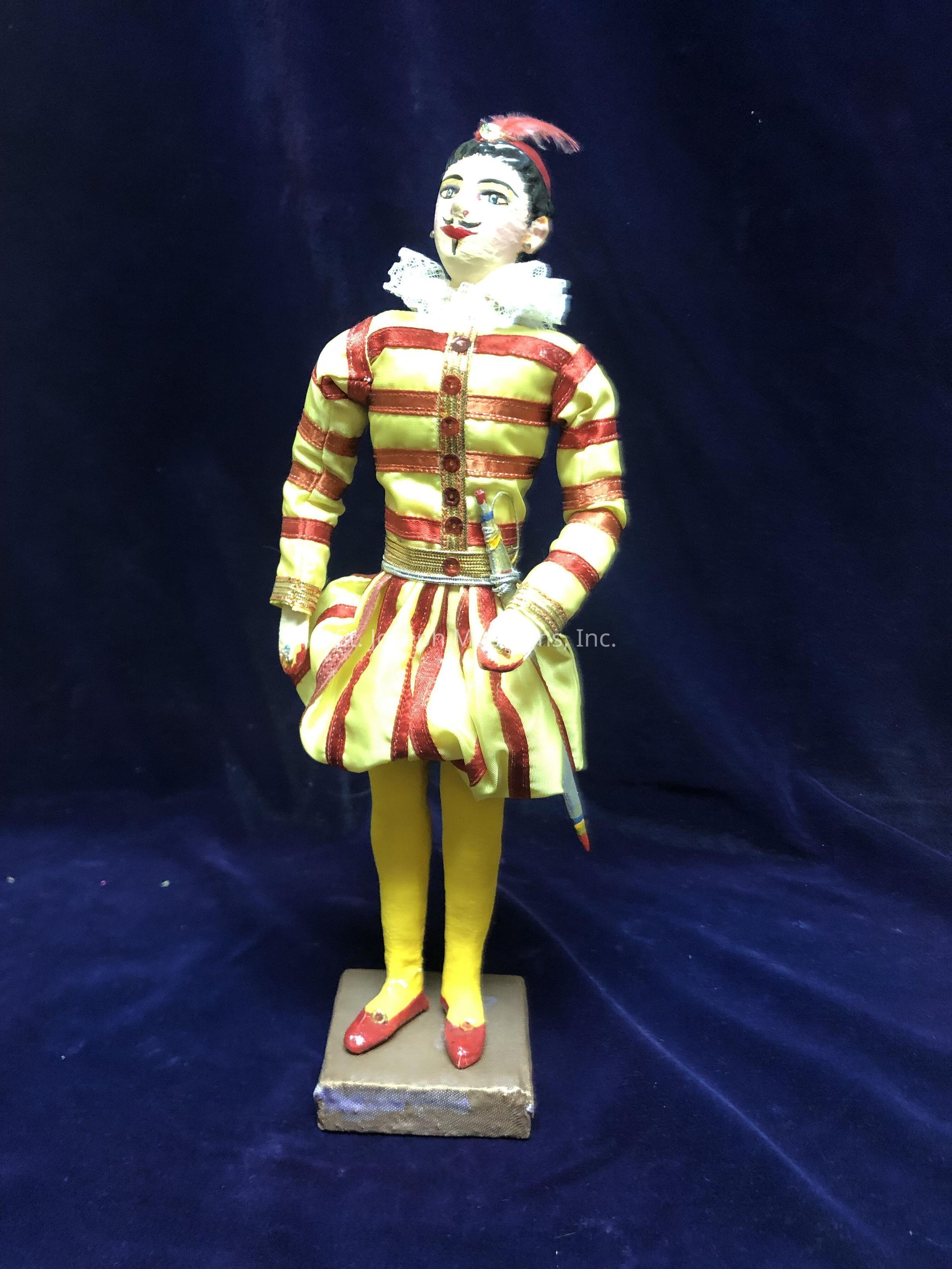 The image depicts a colorful doll dressed in a vibrant, striped costume featuring alternating red and yellow bands. The doll is adorned with a ruffled, white collar, giving it an elegant and theatrical appearance. Its face is painted with distinct features, including bright eyes and red lips, contributing to its expressive look. A small red feather ornament decorates the top of its head, adding to the character's charismatic persona. The doll's legs are covered in bright yellow leggings, and it stands on a rectangular platform, highlighting its neatly crafted red shoes. The background is a deep, rich blue, providing a striking contrast to the vibrant colors of the doll's outfit, further enhancing its visual appeal and theatricality.