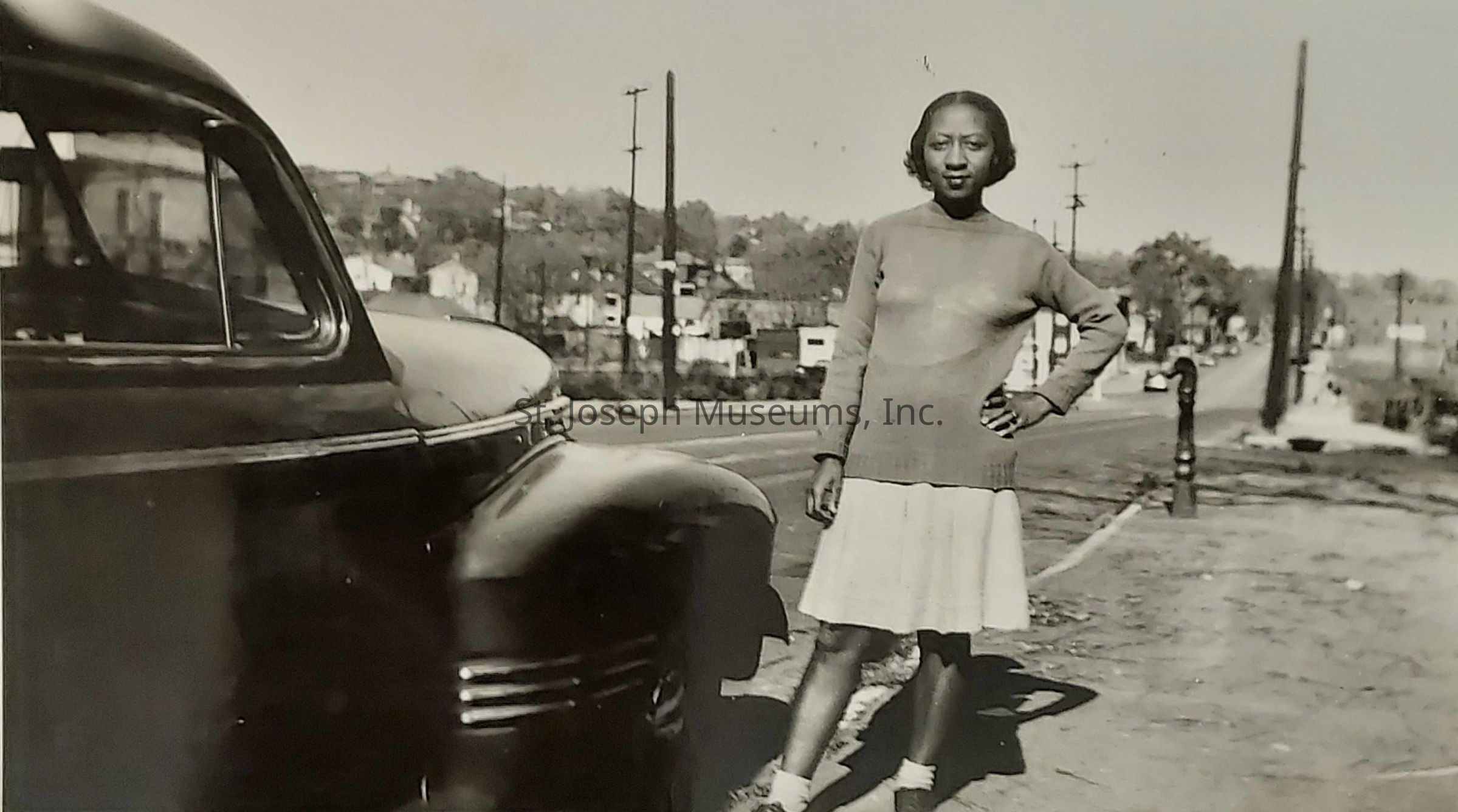 In this black and white photograph, a woman stands confidently with one hand on her hip, dressed in a long-sleeved sweater and a knee-length skirt. She is positioned on a quiet street next to a vintage car, likely from the early to mid-20th century. The background reveals a small town or suburban area with numerous utility poles and modest houses lining the road, suggesting a calm, sunny day. The image captures a moment in time, reflecting the fashion and atmospheric setting of a bygone era, while the woman's relaxed yet self-assured pose adds a personal touch to the historical context.