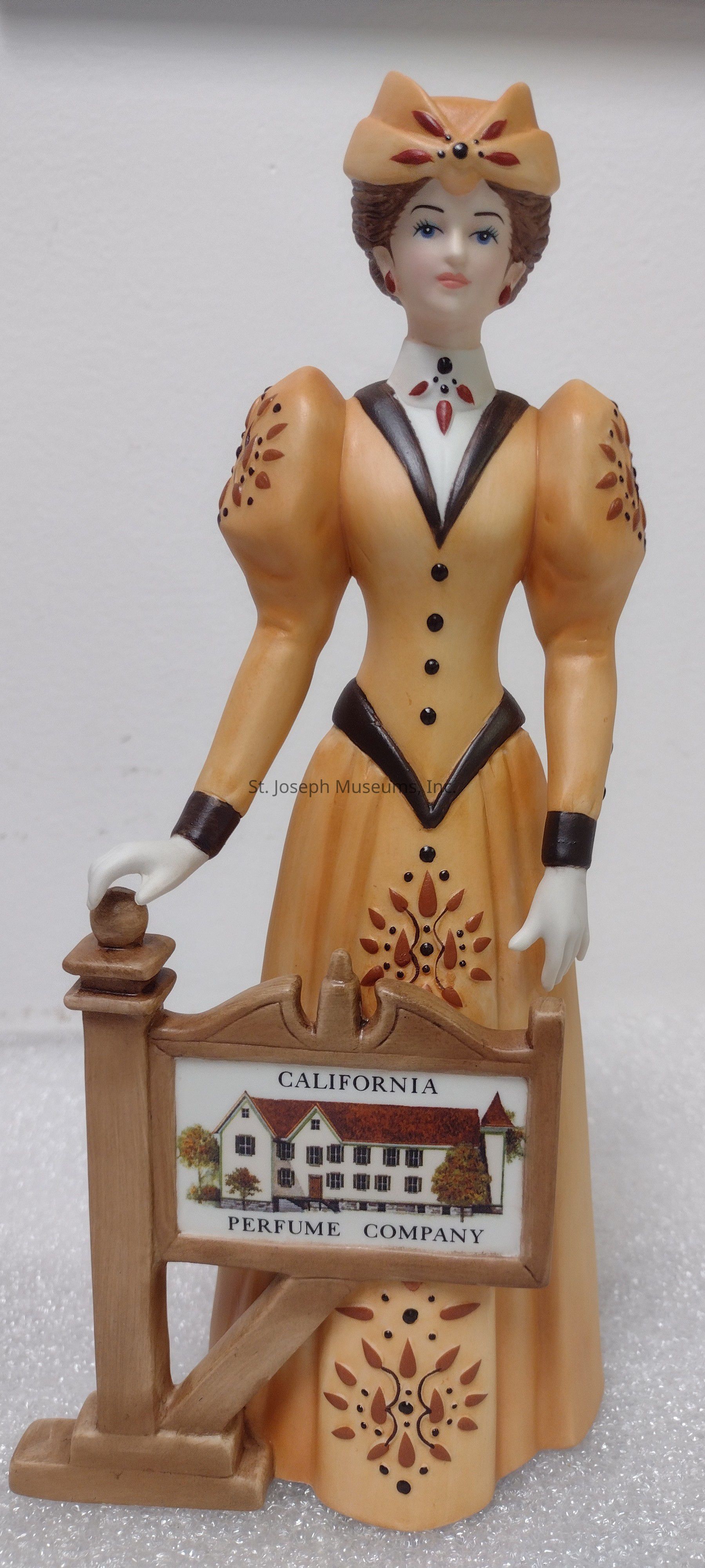 The image depicts a detailed figurine of a woman dressed in vintage clothing, characterized by a light brown long-sleeved dress with intricate decorative patterns and puffed shoulders. She is wearing a matching hat adorned with embellishments and has a calm expression with rosy cheeks and red lips. The woman is holding a wooden frame with a sign that reads "California Perfume Company," featuring an illustration of a large white house with a red roof surrounded by greenery. The figurine stands on a textured surface, capturing the essence of a historical or collectible item from a past era, possibly representing a historical figure or an advertising piece for the company.