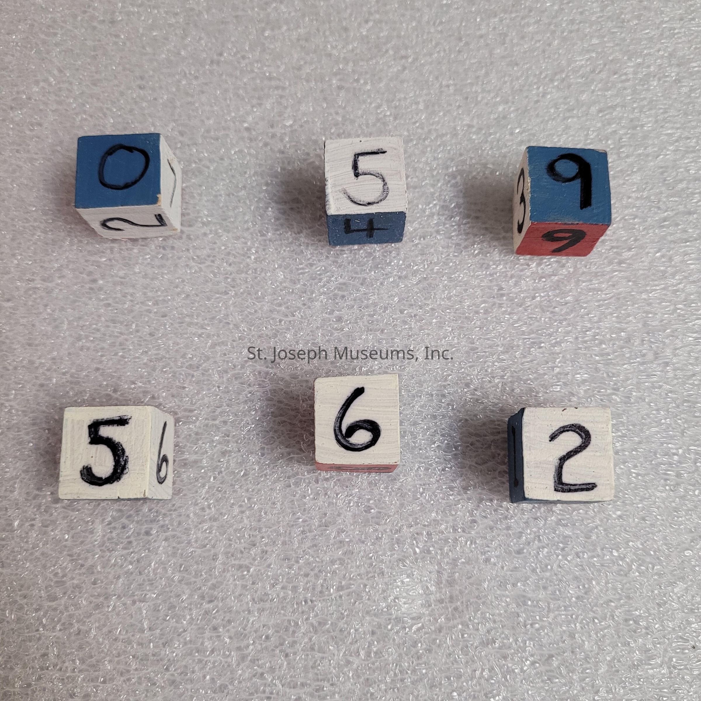 The image shows six wooden cubes, each with different numbers painted on their visible faces. The cubes are arranged in two rows on a textured, white foam surface. In the top row, the cubes display the numbers 0, 5, and 9, with the digits boldly hand-painted in black on a predominantly white and blue background. Below, the second row features the numbers 5, 6, and 2, similarly presented with large black digits contrasting against the light-colored surface of each cube. Each cube has a unique combination of numbers on its various faces, emphasizing a playful and educational design.