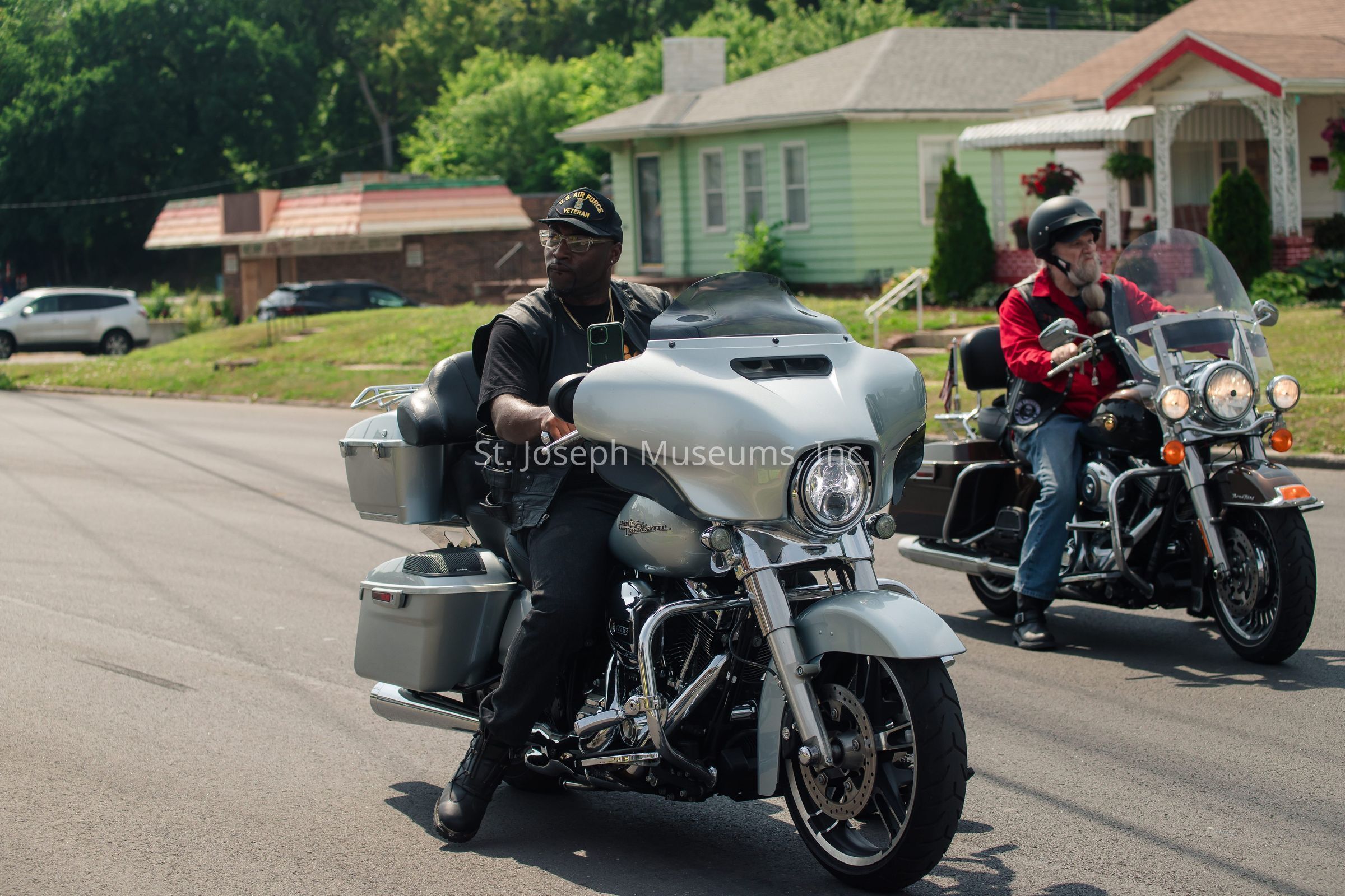 In the image, two individuals are riding motorcycles on a sunny day along a quiet suburban street. The person in the foreground is on a sleek, silver motorcycle, wearing a black cap and a leather vest, appearing focused on the road ahead. To the right, slightly behind, another person on a black motorcycle is wearing a red jacket and a black helmet, maintaining a steady pace. The backdrop features a couple of quaint houses with well-kept lawns, surrounded by lush greenery and a parked car in the distance, suggesting a peaceful neighborhood. The atmosphere conveys freedom and camaraderie as they cruise together under a clear blue sky.