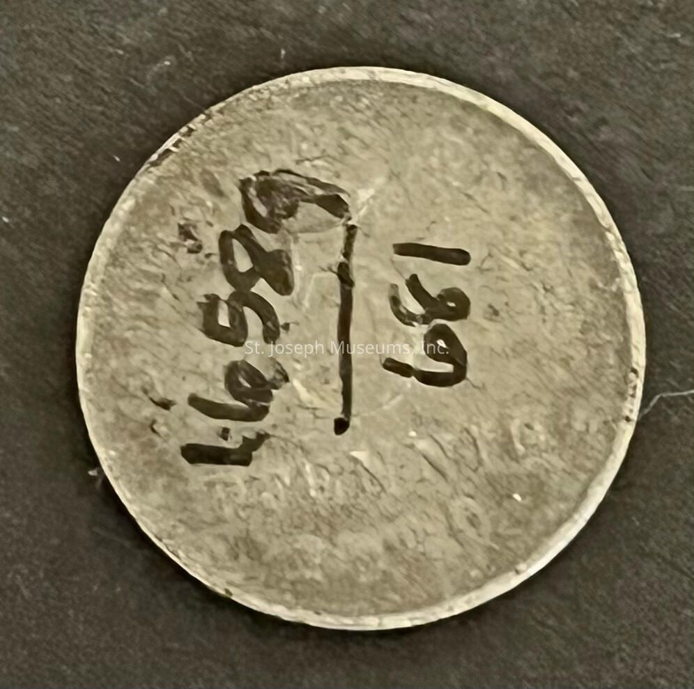 The image depicts a circular metallic object resembling a coin, set against a dark background. The coin has a textured surface with visible scratches and imperfections, suggesting age or use. The handwriting is somewhat crude and irregular, which may imply it was done manually rather than by a machine. The edges of the coin are slightly worn, hinting at handling or exposure to elements over time. The lighting is focused, creating shadows that enhance the texture, while the overall color of the coin is a dull silver or gray. Shadows and highlights contribute to a three-dimensional appearance, emphasizing the roughness and worn condition. This image might represent a token, an identification piece, or simply an artifact with historical or personal significance. The overall impression evokes curiosity about the coin's origin, purpose, and the meaning behind the inscribed numbers.