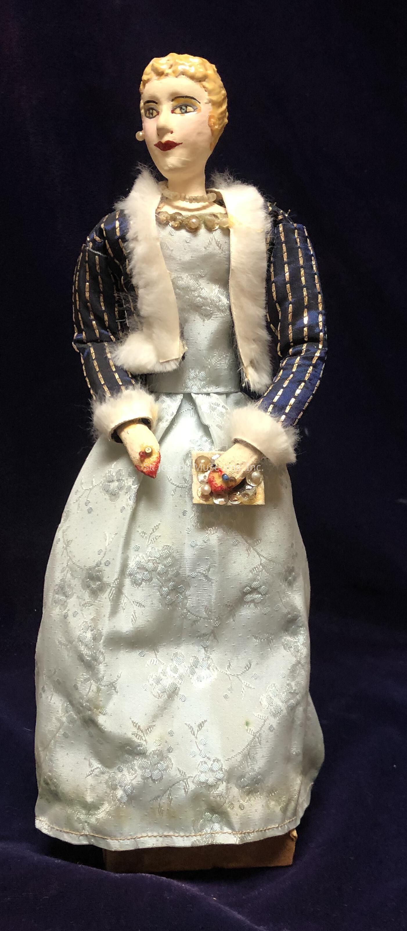 The image depicts an elegantly dressed doll, featuring a detailed face with painted features. She wears a long, light blue gown adorned with subtle floral patterns, conveying an air of sophistication. Over her dress, there is a dark blue jacket with white fur trim and golden stripes, adding a touch of luxury. Her hair is styled in a classic updo, and she holds a small handbag embellished with beads in one hand. The overall appearance suggests a vintage or historical theme, characterized by refined attire and intricate craftsmanship. The background is a deep, dark, indistinct color, which enhances the doll's elegant presentation.