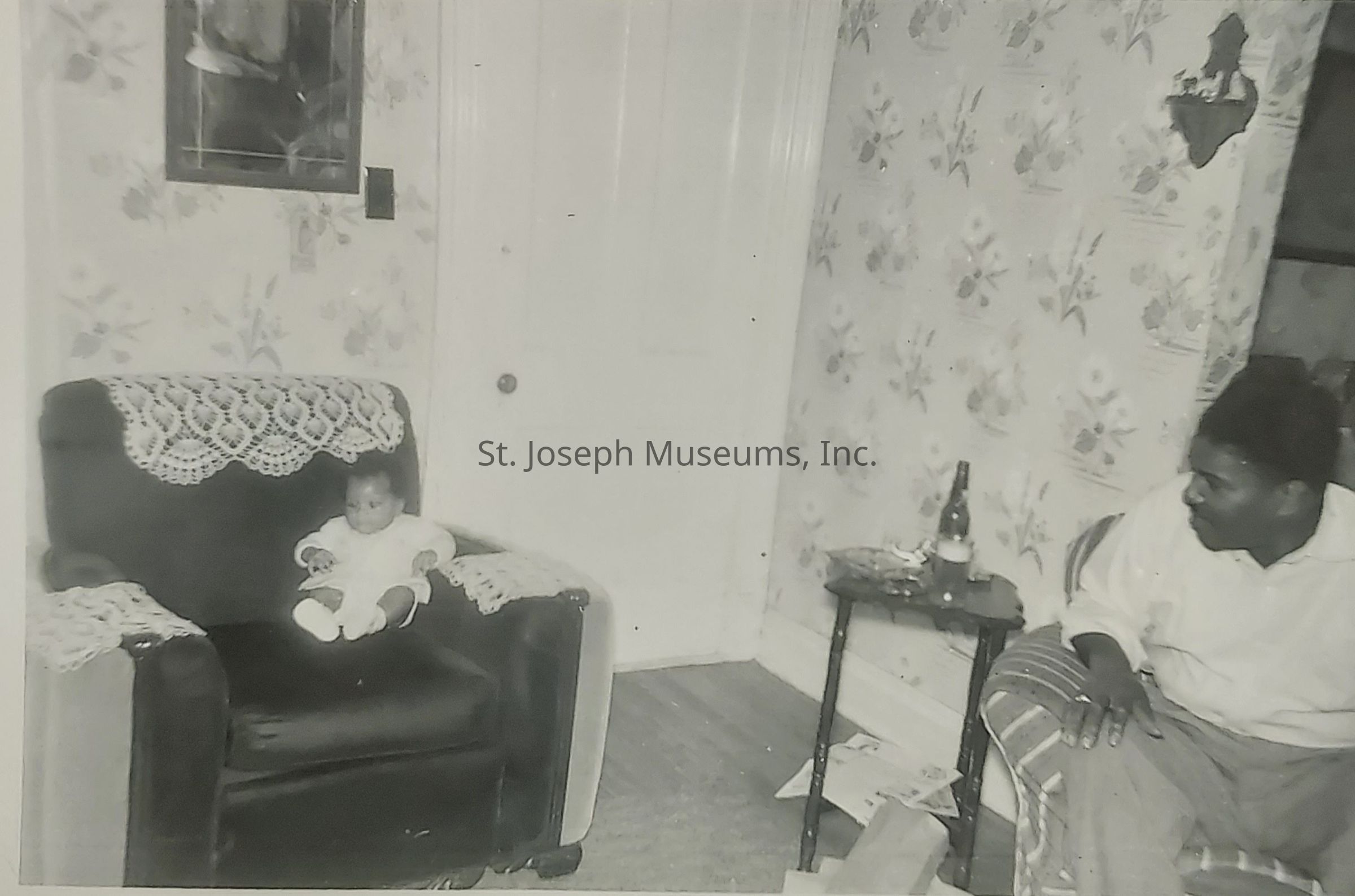 In a cozy, vintage setting, a baby dressed in light clothing is comfortably seated on a large armchair adorned with lace doilies. Nearby, an adult sits on a chair to the right, gazing at the child with interest and affection. The room features floral-patterned wallpaper, blending a sense of warmth and nostalgia. For added character, a small table beside the adult holds a bottle and some scattered papers on the floor, suggesting a lived-in, homely environment. A picture or mirror hangs on the wall, contributing to the vintage ambiance of the scene. The door in the background adds depth to this intimate setting, capturing a moment shared between the two individuals.