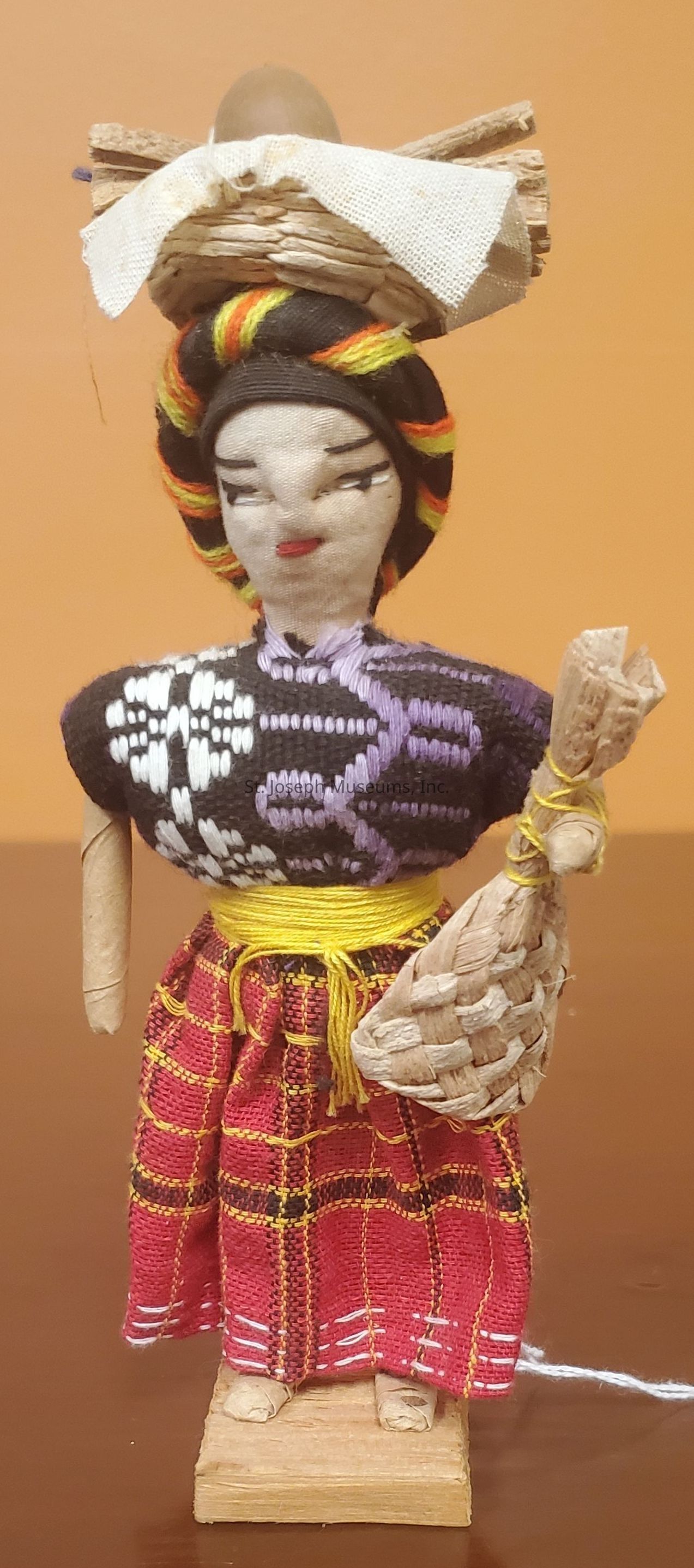 The image shows a handcrafted doll made of various materials, standing on a wooden base. The doll wears a colorful woven outfit, consisting of a black top with white and purple patterns and a red skirt with yellow and black stripes. Its hair is styled with vibrant strands of yellow, orange, and black, and it carries a basket on its head filled with a fabric-covered item and sticks. In one hand, the doll holds a woven bag, and a yellow cord is wrapped around its waist. The overall appearance reflects a traditional or cultural attire theme.