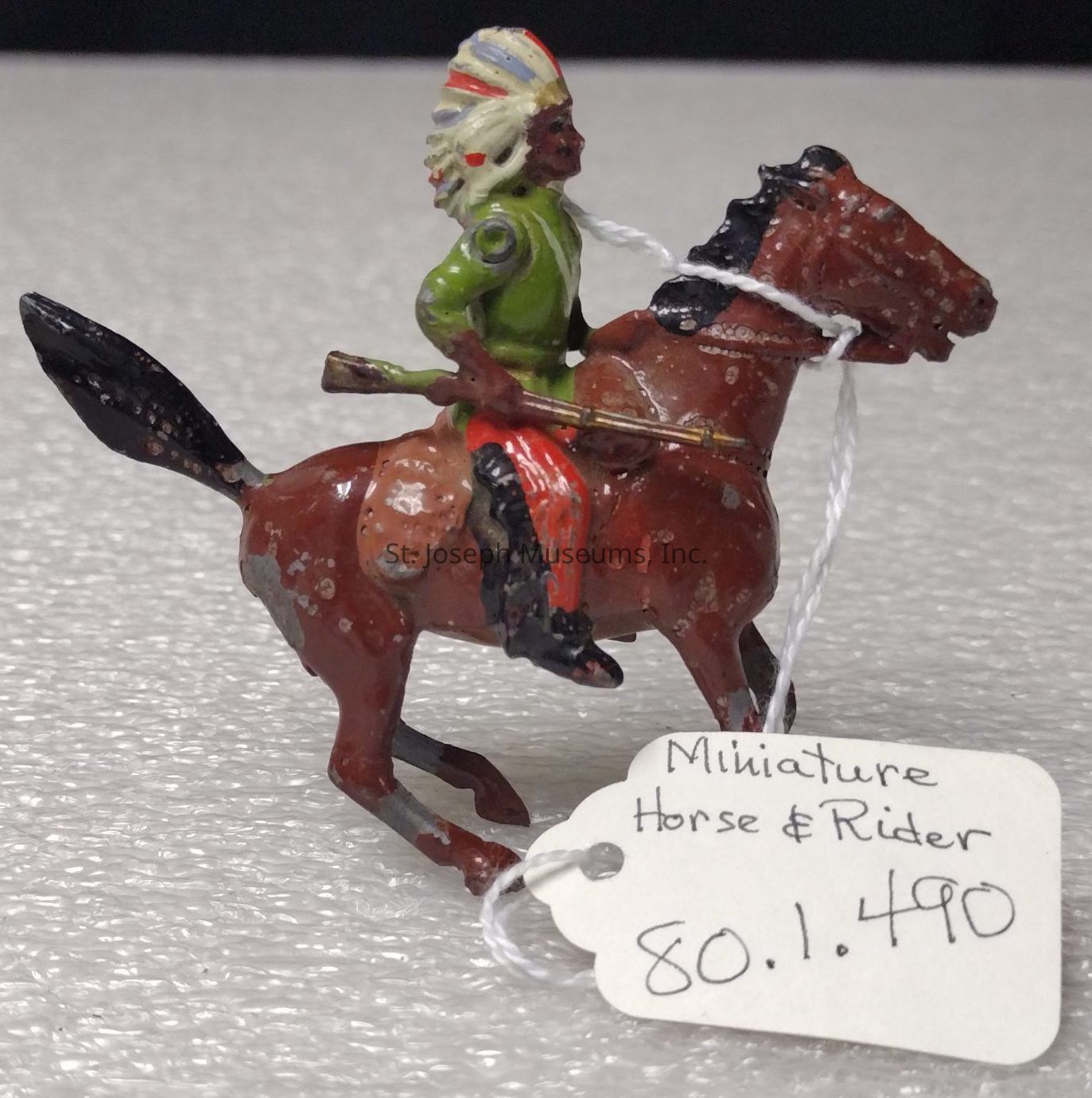 The image shows a miniature figurine of a rider on a horse, crafted with intricate details. The rider is adorned with a colorful headdress and is seated on the brown horse, which is depicted in motion, evident from its raised legs suggesting a gallop. The rider's attire includes a green jacket and red trousers, and they hold an object resembling a rifle in their right hand. A string is tied around the horse’s neck, attached to a white tag labeled “Miniature Horse & Rider” with the number “80.1.490” written below. The figurine features some wear, with visible paint chipping, indicating its age or frequent handling over time. The background is a simple textured gray surface, allowing full focus on the intricacies and vibrant colors of the figurine. This small sculptural piece likely serves as a collectible or display item, capturing the essence of a dynamic horseback scene in a compact form.