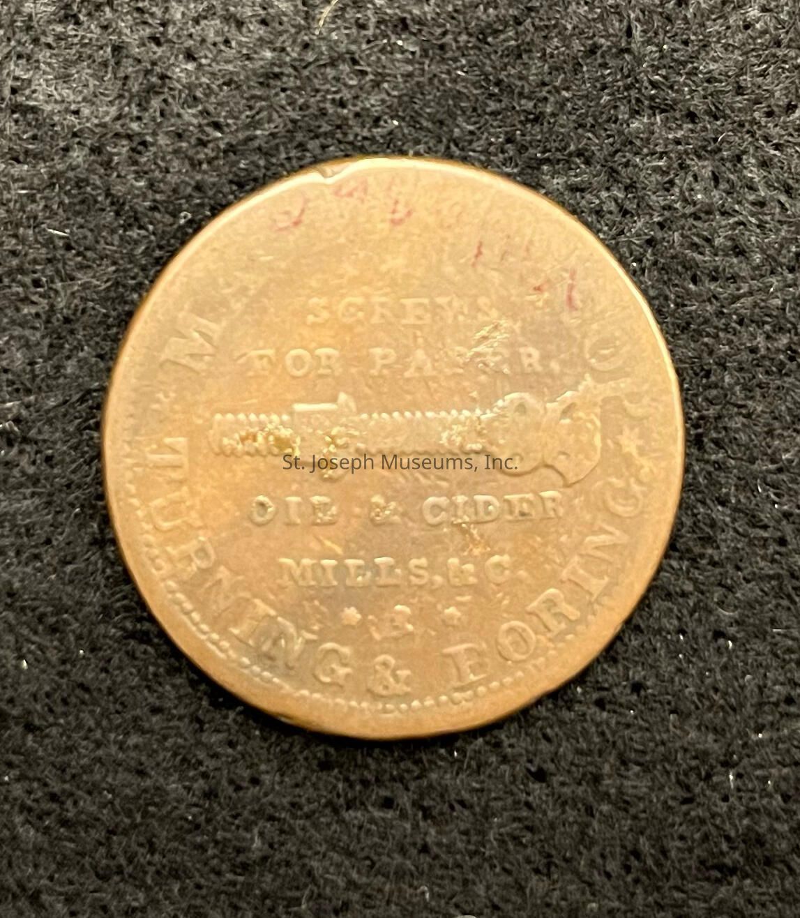 The image shows a close-up of a vintage token or coin placed against a textured black surface. The coin is bronze in color and features detailed engravings, including the words “SCREWS" and "OIL & CIDER" along with “TURNING & BORING,” indicating some historical context or purpose, possibly related to an industrial or commercial activity. The token also displays an image of a screw, further suggesting its connection to machinery or manufacturing. The coin's edges are slightly worn, which might imply age or extensive handling, adding a layer of historical intrigue to the piece.