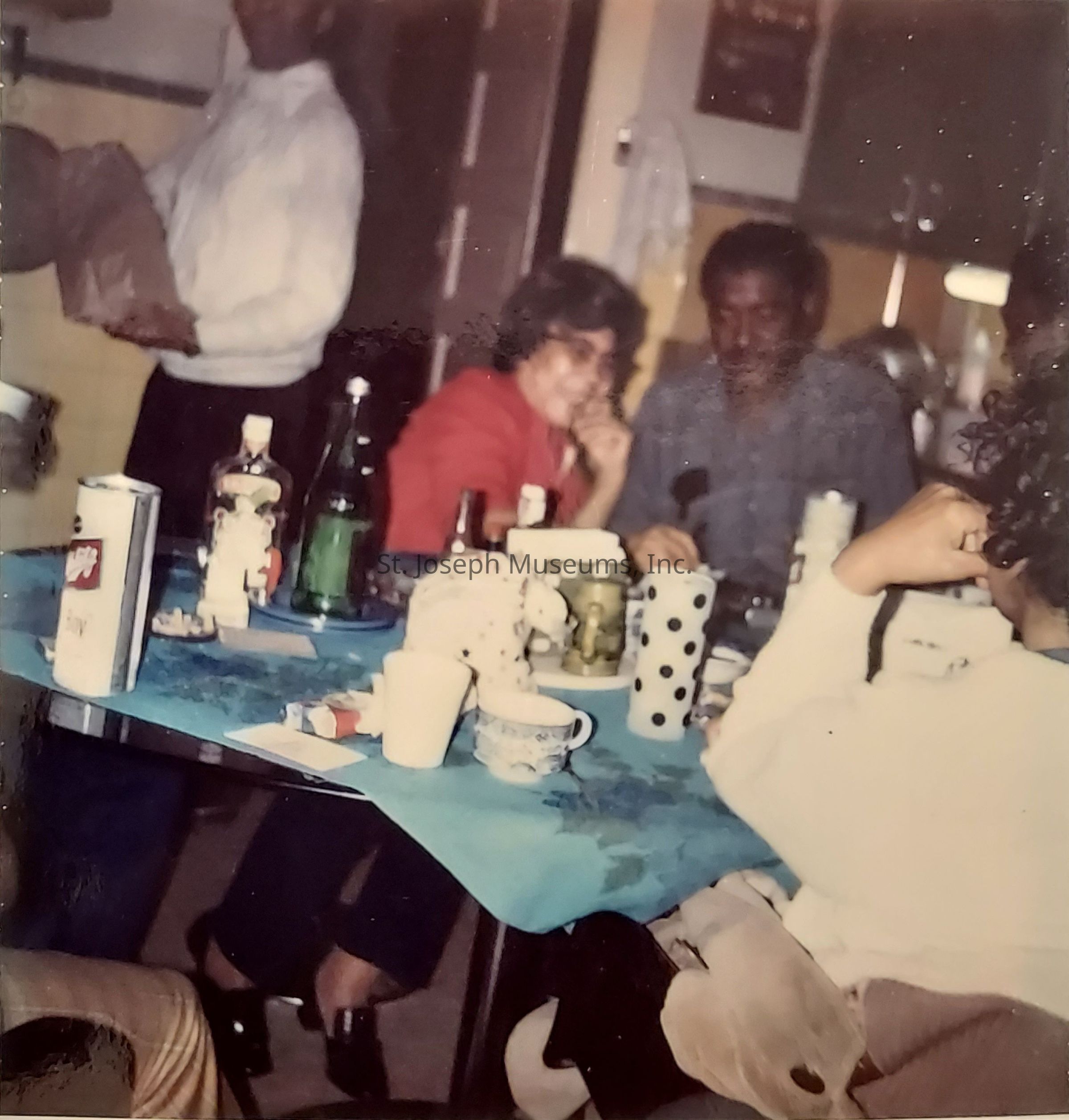 The image depicts a cozy indoor gathering where several people are seated around a table covered with a blue tablecloth. On the table, there are a variety of items including a few bottles, cups with different designs, and other miscellaneous objects such as a stuffed toy. The people appear to be engaged in conversation or simply enjoying each other's company, with one individual wearing a red jacket and smiling, while others sit around the table, visibly relaxed in casual attire. In the background, a person is standing, perhaps holding a brown paper bag, which adds to the informal, homely atmosphere of the scene. The setting suggests a warm and friendly social environment, typical of a small family or friends gathering, possibly during a meal or a casual meet-up. The room appears to be a kitchen or dining area, indicated by the presence of cabinets and various kitchen appliances and containers visible in the background, contributing to the welcoming and informal ambiance.