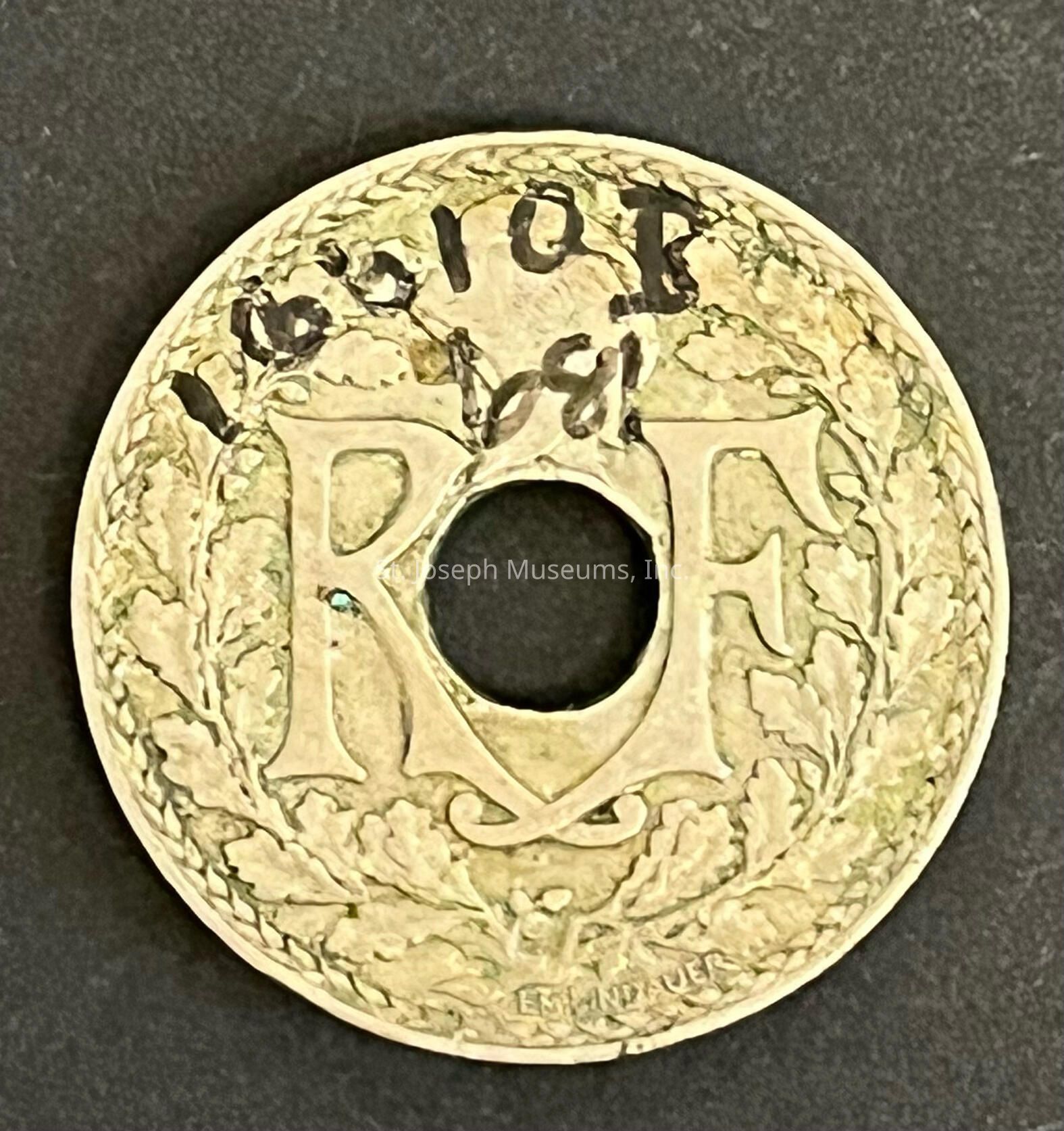 The image depicts an old coin with a circular hole in the center, featuring prominent inscriptions "RF" surrounded by an intricate design of leaves, likely oak or laurel, which is etched around the perimeter. The coin appears to be worn, indicative of its age. The background on which the coin is placed is a plain, dark surface. The coin’s design suggests it might be of French origin, as "RF" could denote République Française. This piece of numismatic history not only tells a story of its own but also showcases classic coin artistry, with every detail—from the foliage design to the handwritten ink—hinting at its journey through time.