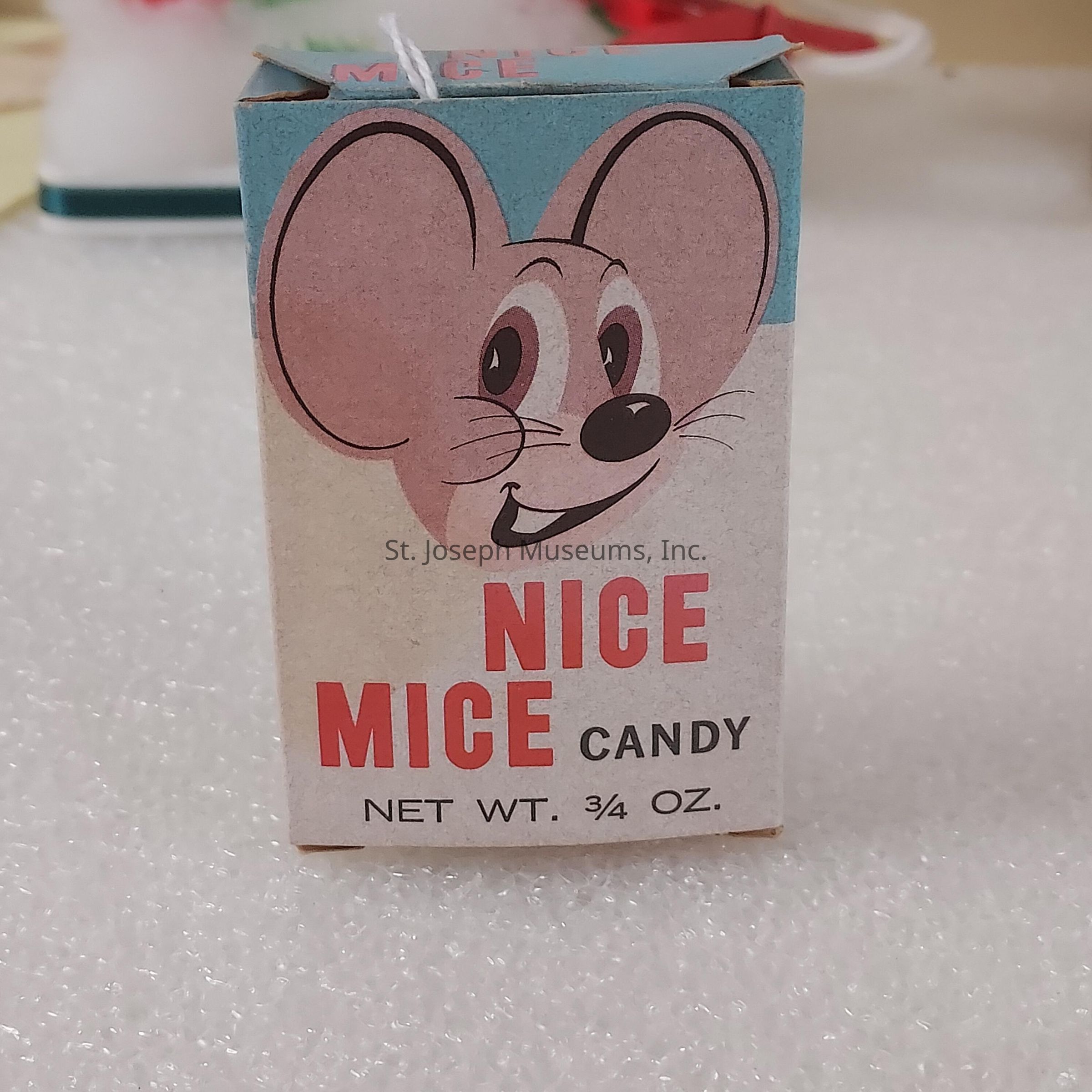 The image shows a small, vintage-style candy box with a whimsical illustration of a smiling cartoon mouse, featuring large, round ears and a friendly expression. Above the mouse, the background is a soft blue, while the box itself is predominantly beige and has bold red lettering that says "NICE MICE CANDY." Additionally, the net weight of the candy is specified as three-fourths of an ounce. The box's nostalgic design evokes a sense of retro charm, making it look like a delightful collectible that could evoke fond memories of childhood treats. The background is a textured white surface, offering a contrasting backdrop that helps the colorful design elements of the box stand out more prominently. This charming packaging speaks to a bygone era where candy boxes often featured playful and inviting artwork to attract young customers.