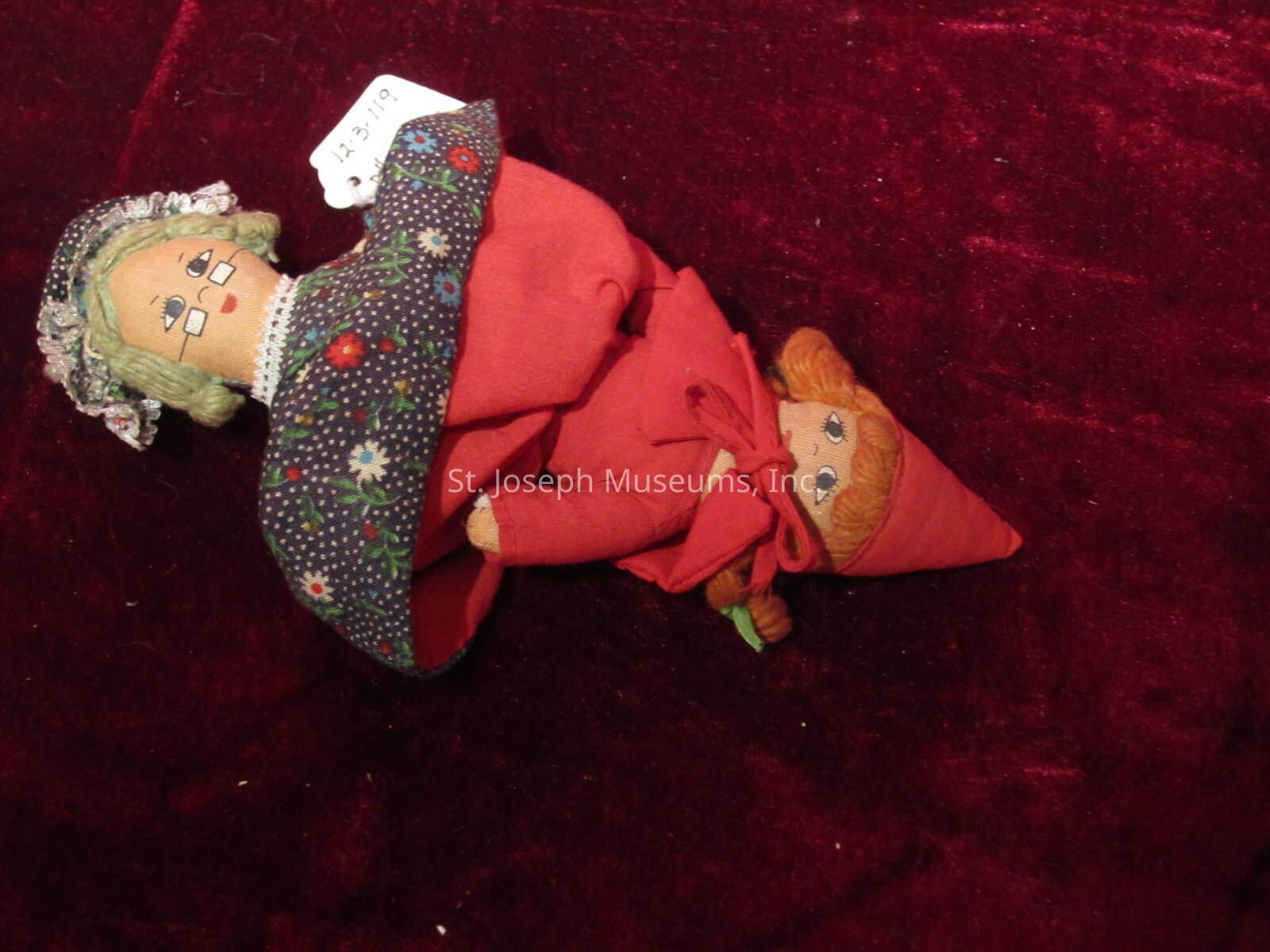 The image features a unique, handcrafted fabric doll that appears to depict two characters in one design, lying on a rich burgundy background. The upper part resembles an elderly character with white hair, round glasses, and a bonnet decorated with an intricate floral pattern against a dark background. This section of the doll is dressed in a red outfit trimmed with lace. Attached to the other side, or perhaps hidden beneath a wrap, is a depiction of a younger character, featuring vibrant red hair and expressive eyes, adorned in a pointed red hat and matching cloak with a bow. A small white price or identification tag marked with a handwritten number is attached to the elderly character's bonnet, suggesting it might be part of a collection or an item for sale. The craftsmanship is detailed, and the dolls appear soft and stitched, displaying a blend of artistic creativity and folk-art style. The red and green color scheme enhances the visual contrast, adding an additional layer of depth and cultural storytelling to the piece.