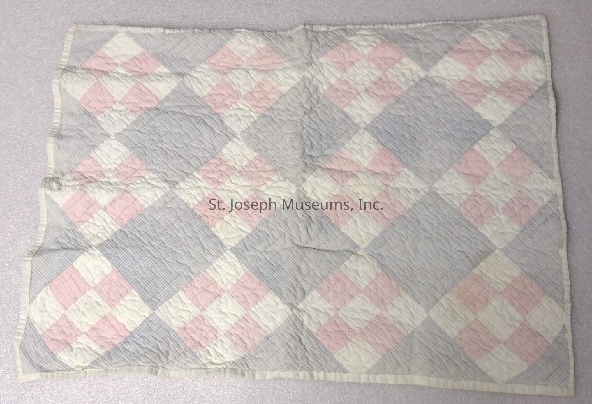 The image features a quilt laid out flat, showcasing a patchwork design made up of pastel colors, specifically pink, white, and gray. The quilt's pattern consists of square blocks arranged diagonally, creating a diamond-like effect. Within each diamond, alternating squares of pink and white are stitched against a subtle gray background, giving the quilt a soft and gentle appearance. The edges of the quilt are bordered with a light-colored fabric, neatly sewn to provide a finished look. The entire quilt exhibits a slightly crinkled texture, adding a sense of coziness and warmth to its overall aesthetic.