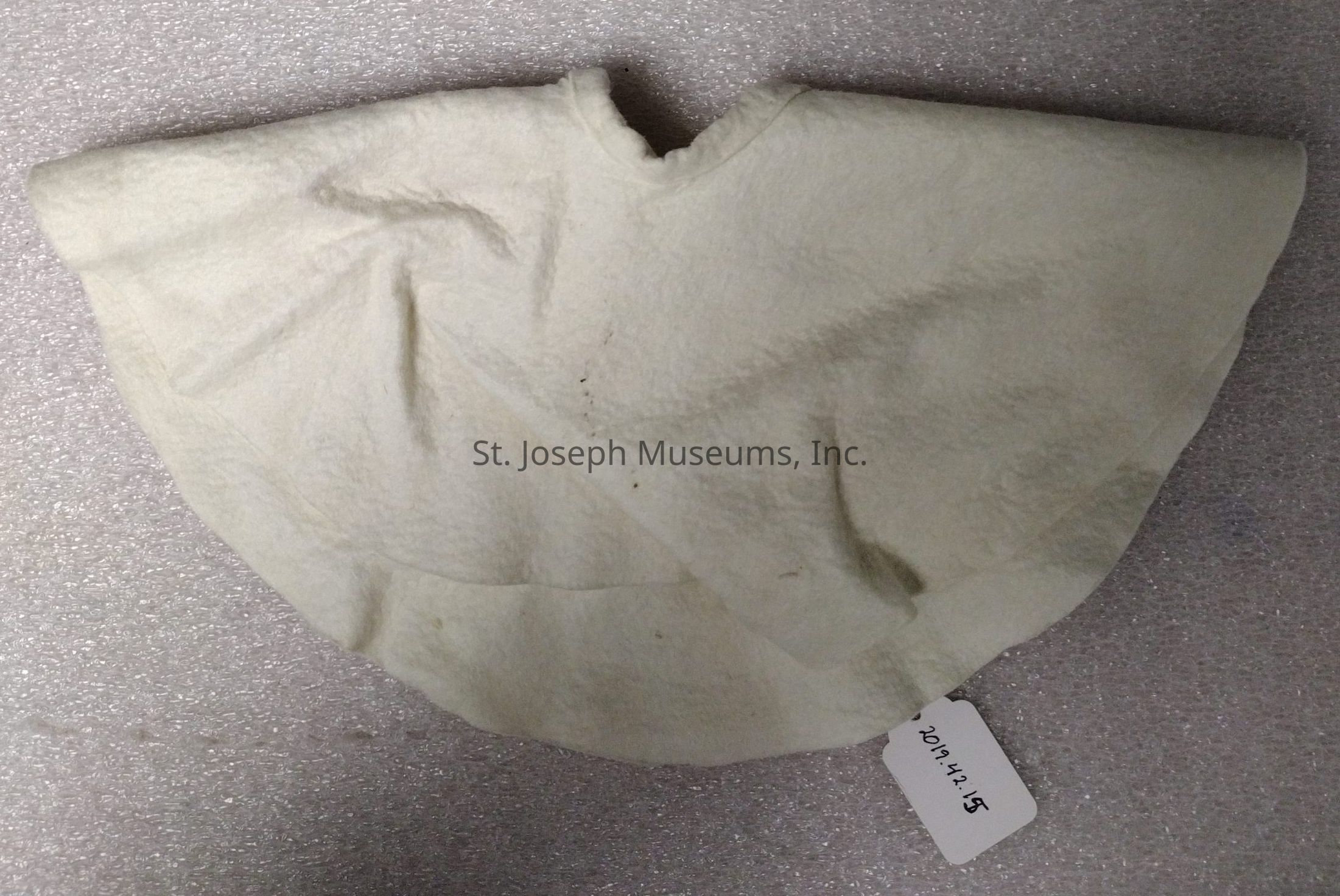 The image depicts a fabric object with an off-white hue, featuring a rounded, slightly pleated shape, possibly indicating its use as a garment or accessory. The fabric's texture appears soft, with light creases suggesting it might have been folded or worn. A small, rectangular white tag is visible, attached at the lower edge, displaying the numerals "2014.1.15" in black ink, which may refer to an identification number or a catalog reference. The background consists of a textured surface resembling speckled, light gray material, providing a neutral backdrop that emphasizes the fabric's relatively smooth appearance.