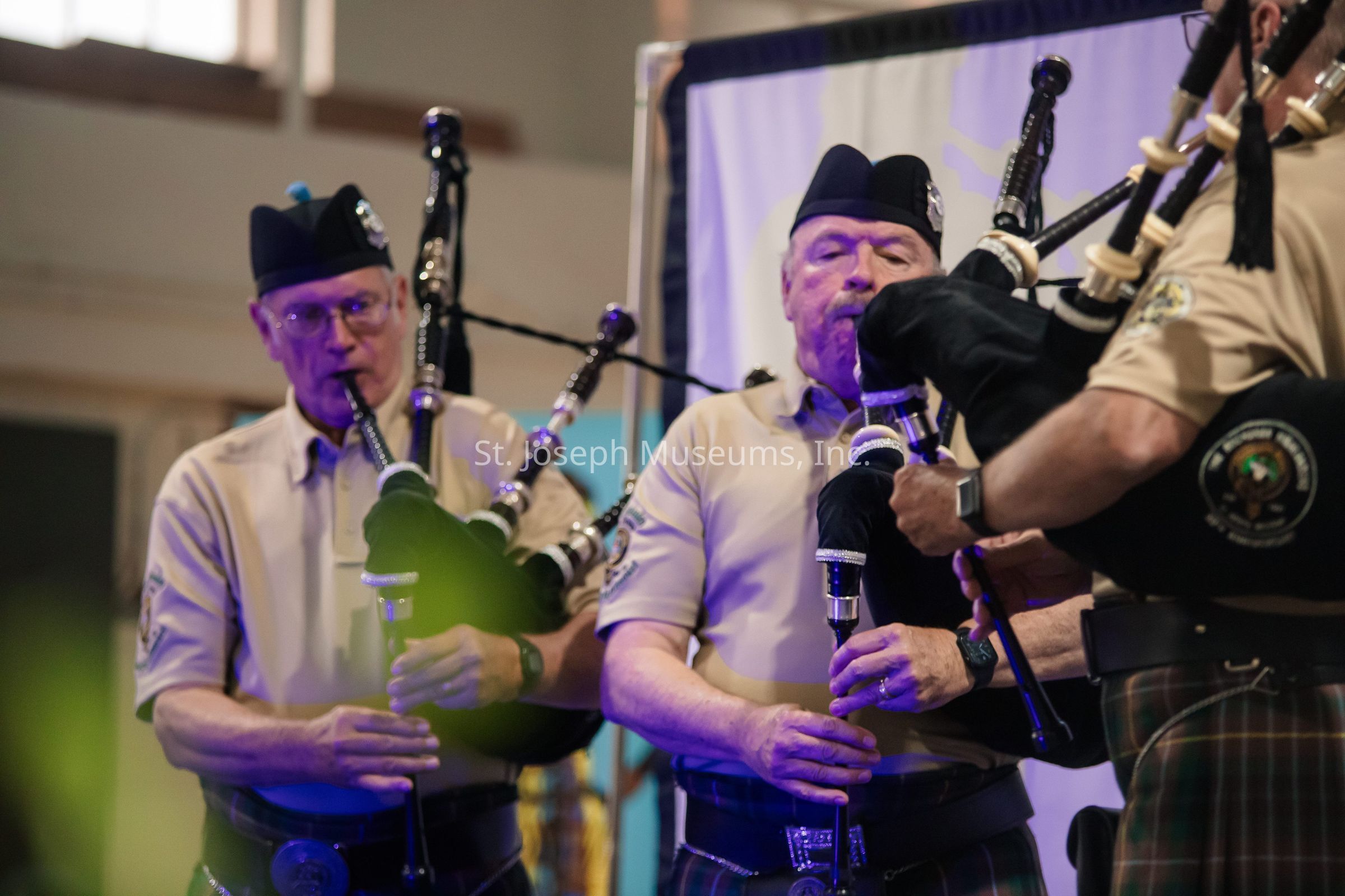 The image captures a group of musicians in a well-lit indoor setting, each intensely focused as they play the bagpipes, wearing coordinating khaki shirts and traditional Scottish kilts, with matching dark caps that feature badges on the front. The musicians are engaged in their performance, with one person in the foreground clearly visible as they blow into the instrument, creating a lively and energetic atmosphere. The bagpipes they hold are adorned with intricate details, and their presence, along with the attire, contributes to a traditional and culturally rich performance setting. In the background, a partially blurred banner hints at the event or gathering type, while soft, warm colors create a welcoming and vibrant ambiance, making the overall scene both dynamic and visually appealing, emphasizing the celebration of heritage through music.