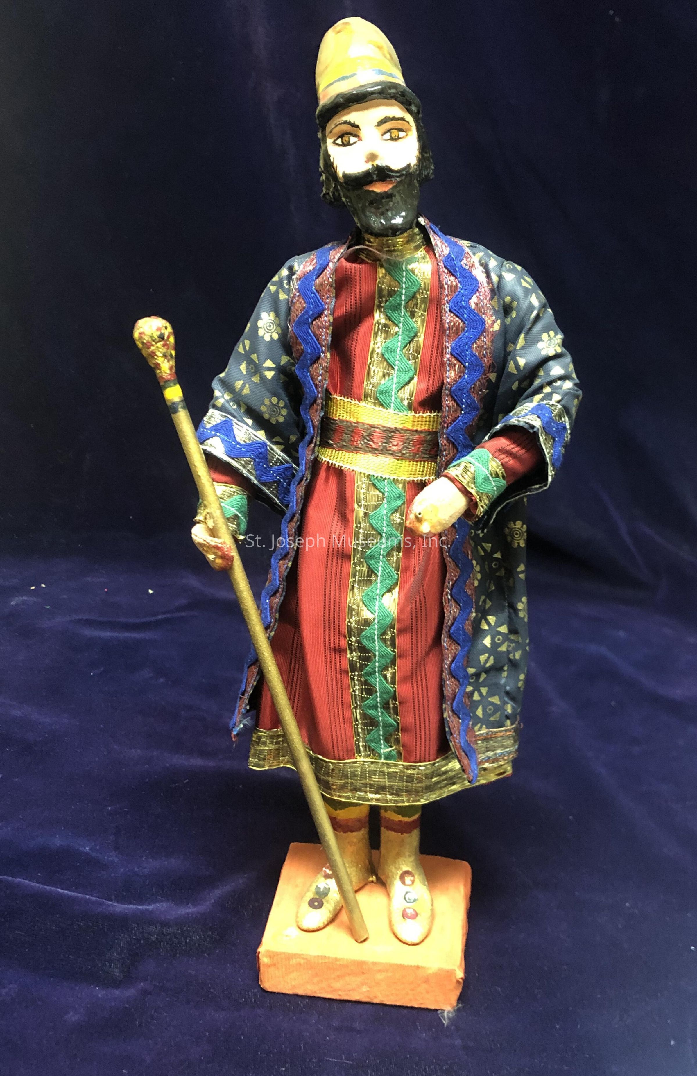 The image shows a handcrafted figurine of a person adorned in traditional attire, featuring a colorful, intricately patterned robe with zigzag and floral designs in shades of blue, red, and gold. The figure wears a tall hat with gold accents, matching the ornate details of their ensemble and is positioned on a simple orange platform. In one hand, the figure holds a decorative staff, adding to the ceremonial feel conveyed by the attire and pose. The face of the figurine is expressive with painted eyes and a prominent mustache and beard, contributing to its lifelike appearance. The background is a deep, rich shade of blue, which contrasts and enhances the vibrant colors of the figure's clothing, highlighting the artisan's attention to detail and cultural influences in the design. The overall craftsmanship reflects a blend of cultural artistry and traditional fashion elements, capturing a sense of elegance and historical significance.