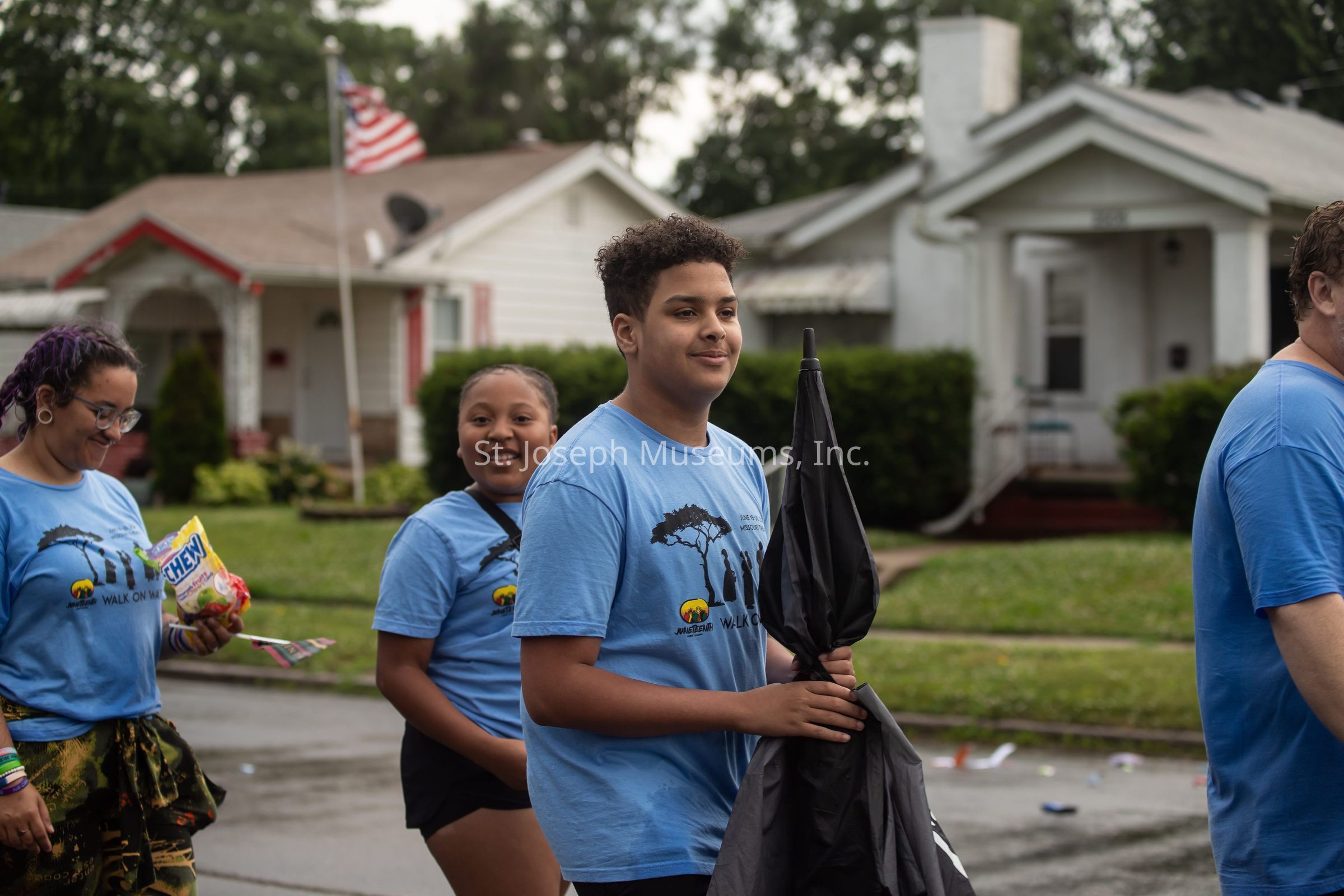 The image depicts a group of people participating in an outdoor event, all wearing matching light blue shirts with black designs indicating a theme of unity or a specific cause. In the foreground, a young person with curly hair holds a large, folded black umbrella and smiles softly, appearing to be part of a casual, community gathering or parade. Beside them, a girl with braided hair and a small backpack smiles cheerfully at the camera, suggesting an atmosphere of friendliness and community spirit. Another person, with brightly colored hair, looks down while holding a bag of snacks, perhaps preparing to share them. The background shows neat, suburban houses with manicured lawns, and an American flag is visibly flying atop a flagpole, hinting at a national holiday or local celebration. The scene conveys a sense of togetherness and shared purpose, as the group walks along a street with scattered bits of debris or decorations on the ground, against a backdrop of lush green bushes and trees, suggesting a recent event or ongoing celebration.