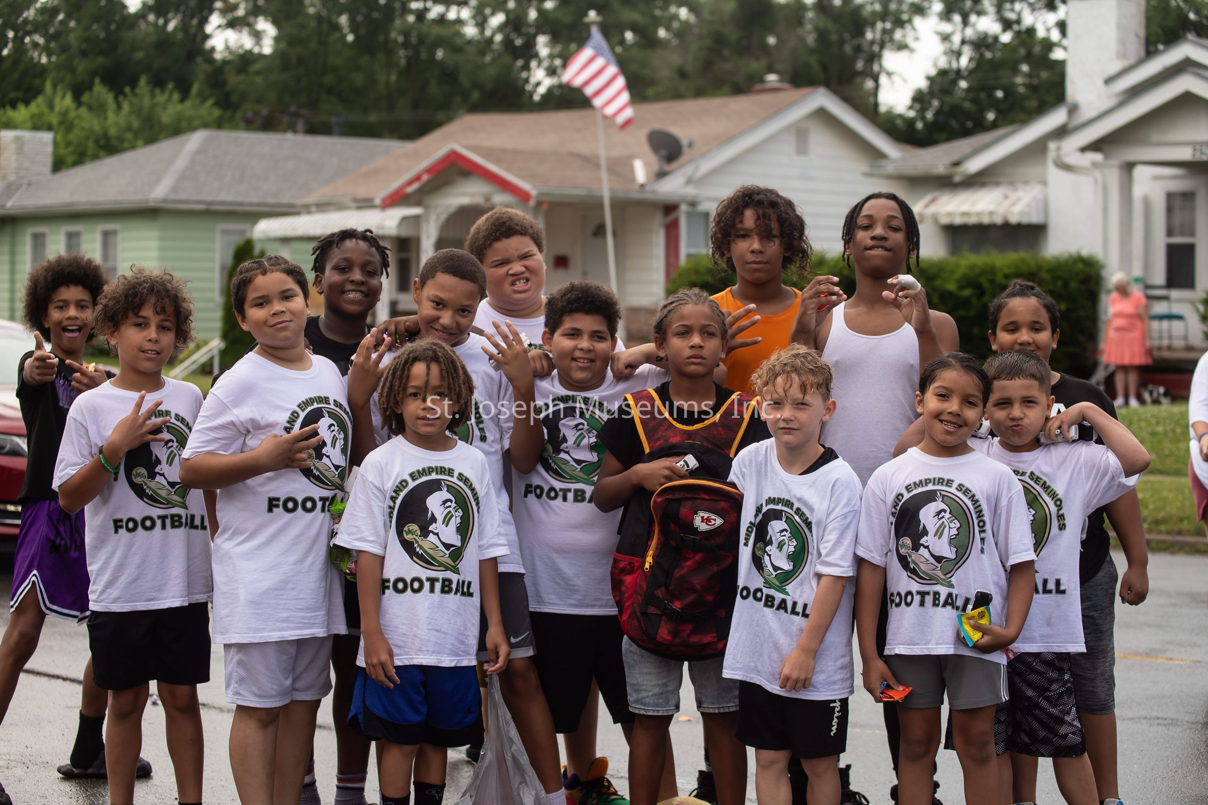 The image depicts a group of young boys and girls standing together on a residential street, all wearing matching T-shirts that say "Inland Empire Seminoles Football," indicating they are part of a football team, and they pose joyfully with various hand gestures, while behind them are houses with a visible American flag proudly flying against a backdrop of trees and a cloudy sky.