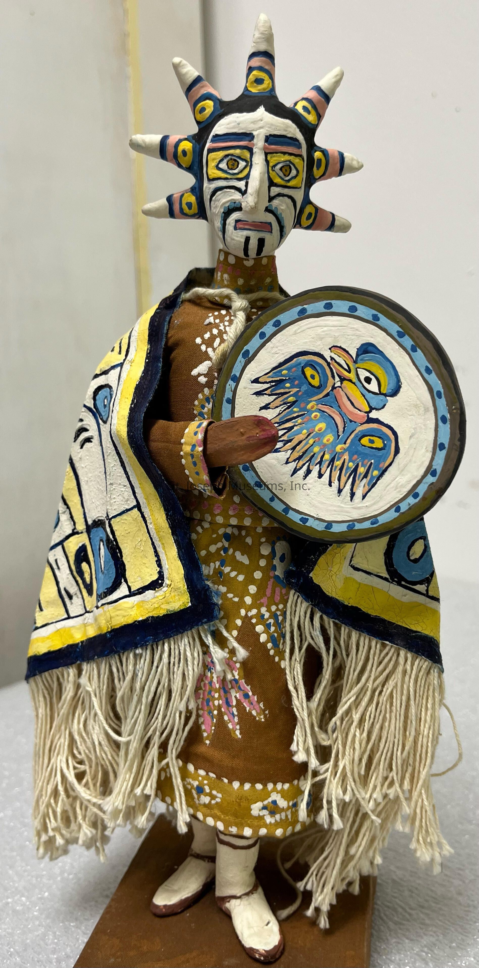 The image depicts a handcrafted figurine with an intricate, colorful design, featuring a human-like form adorned with a traditional mask resembling a sun with pointed rays, painted in vivid hues of blue, yellow, and black. The figure wears an elaborately patterned brown garment decorated with white and pink dots and motifs, and a fringed cloak in shades of yellow and blue, accentuating the cultural artistry. It holds a round shield or drum, adorned with a painted bird design in blue and yellow, complementing the overall aesthetic and implying a ceremonial or cultural significance to the piece. The craftsmanship suggests a representation of heritage or storytelling, with detailed attention to balance and vibrant coloration. The figure stands on a brown base, completing the presentation, showcasing not just an item but a unique embodiment of artisanal creativity and tradition, possibly reflecting indigenous or folk influences in its style and symbolism. This artistic piece, with its combination of materials and decorative elements, brings life to cultural narratives.