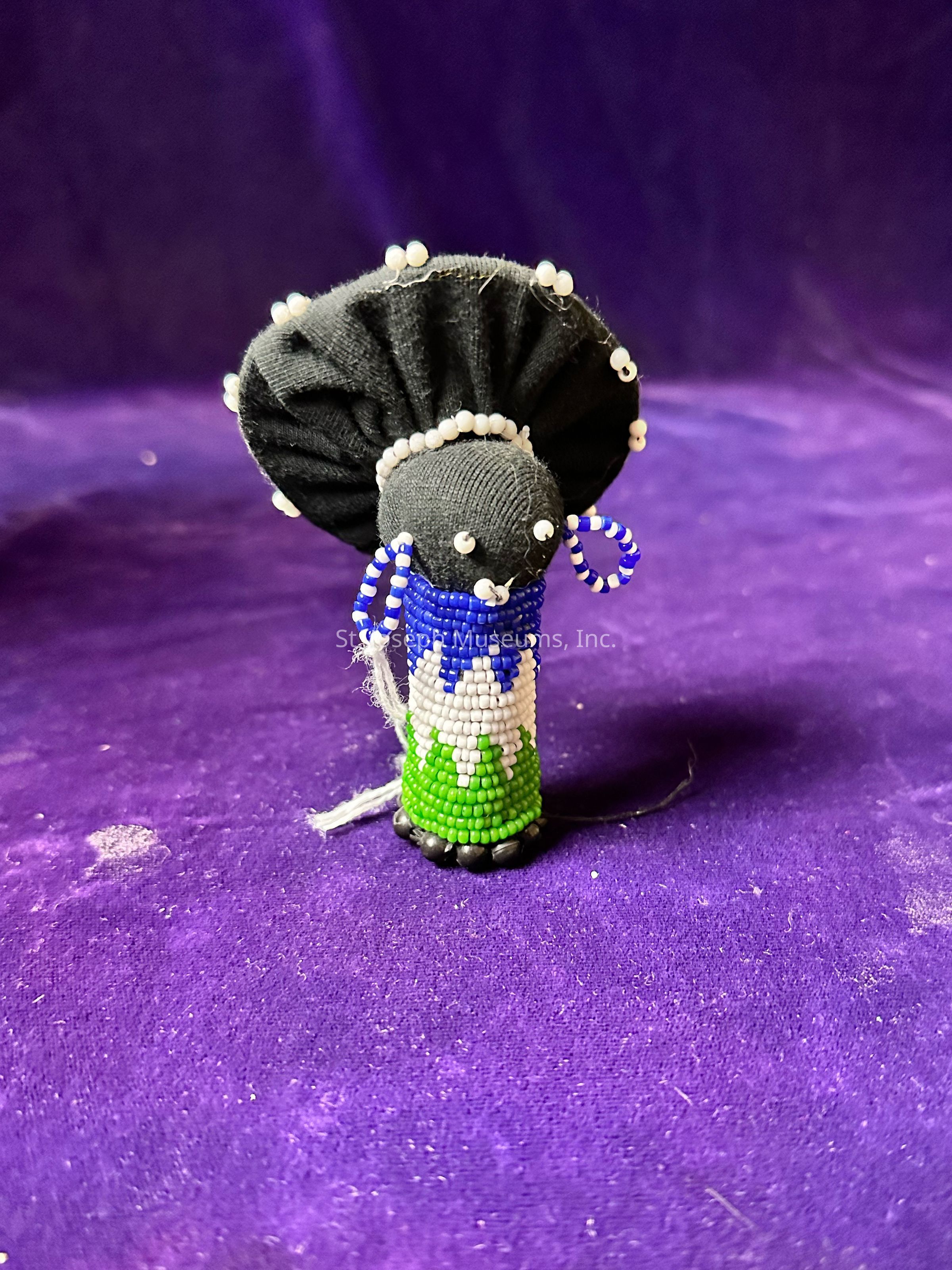 The image showcases a small, handcrafted figure set against a rich purple background. The figure is adorned with intricate beadwork, featuring a vibrant combination of blue, white, and green beads forming its body. Its head is covered with a black fabric that is elegantly gathered at the top, embellished with tiny white beads that accentuate the design. The figure appears to have small bead-crafted eyes and wears a pair of beaded earrings, adding to its detailed craftsmanship. The overall texture and color contrast beautifully with the deep purple backdrop, highlighting the artistry and cultural essence of the figure.